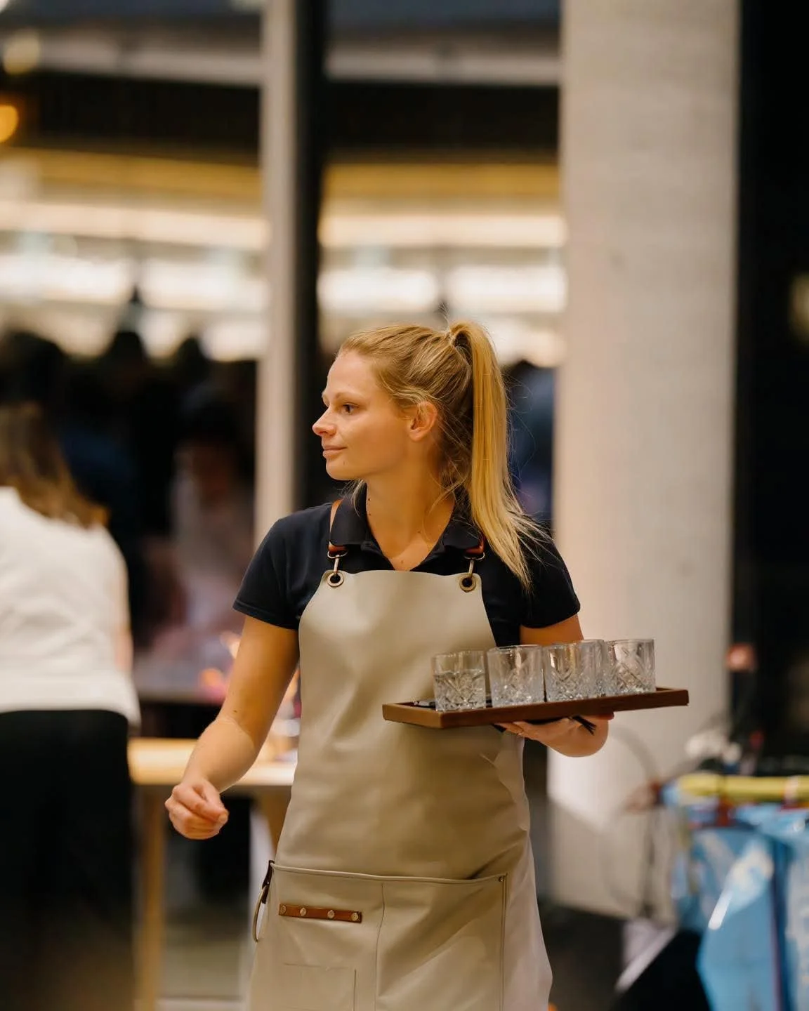 Femme portant un tablier beige, tenant un plateau avec des verres dans un environnement intérieur, probablement un restaurant ou un café.