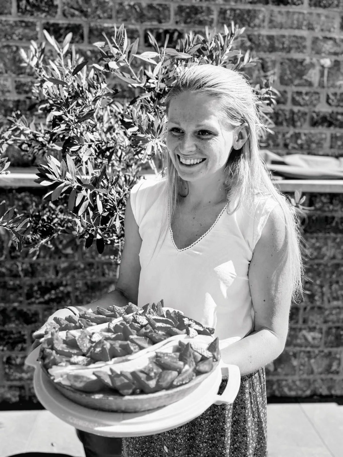 Femme souriante tenant un plateau de pâtisseries à l'extérieur, avec un mur en briques en arrière-plan.