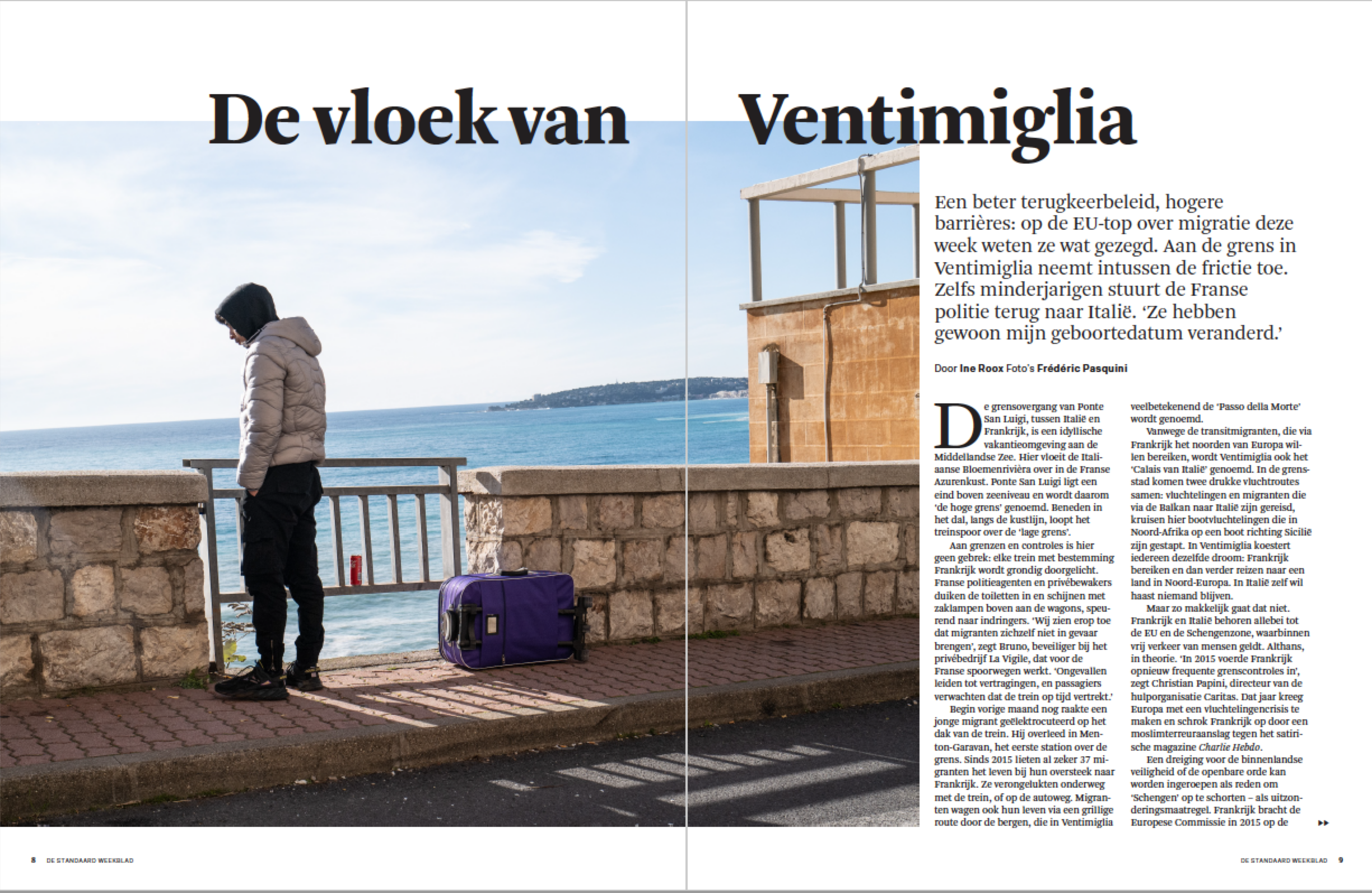 A person standing by a seaside terrace, wearing a beige puffer jacket and black pants, with a purple luggage bag beside them. The person is looking down with their hood up, near a stone wall with the ocean and horizon in the background.