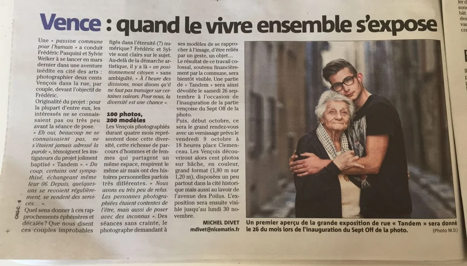 Newspaper article with a photo of a young man and an elderly woman embracing outdoors. The young man is wearing glasses and a dark t-shirt, and the elderly woman has white hair and is wearing a dark sweater with a polka dot collar. The article headli