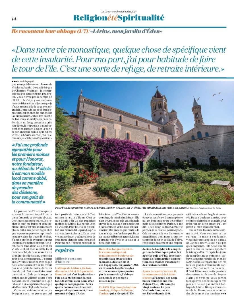 Photograph of Lerins Abbey in Lyon, France, with stone walls, towers, and vibrant purple flowers in the foreground, under a clear blue sky with a few birds flying.