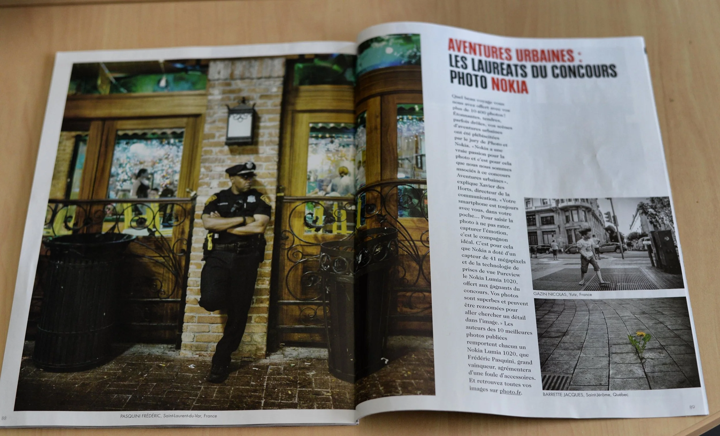 A police officer leaning against a brick and wood building, with a decorative iron fence in front, seen in a magazine spread about urban photography competitions.