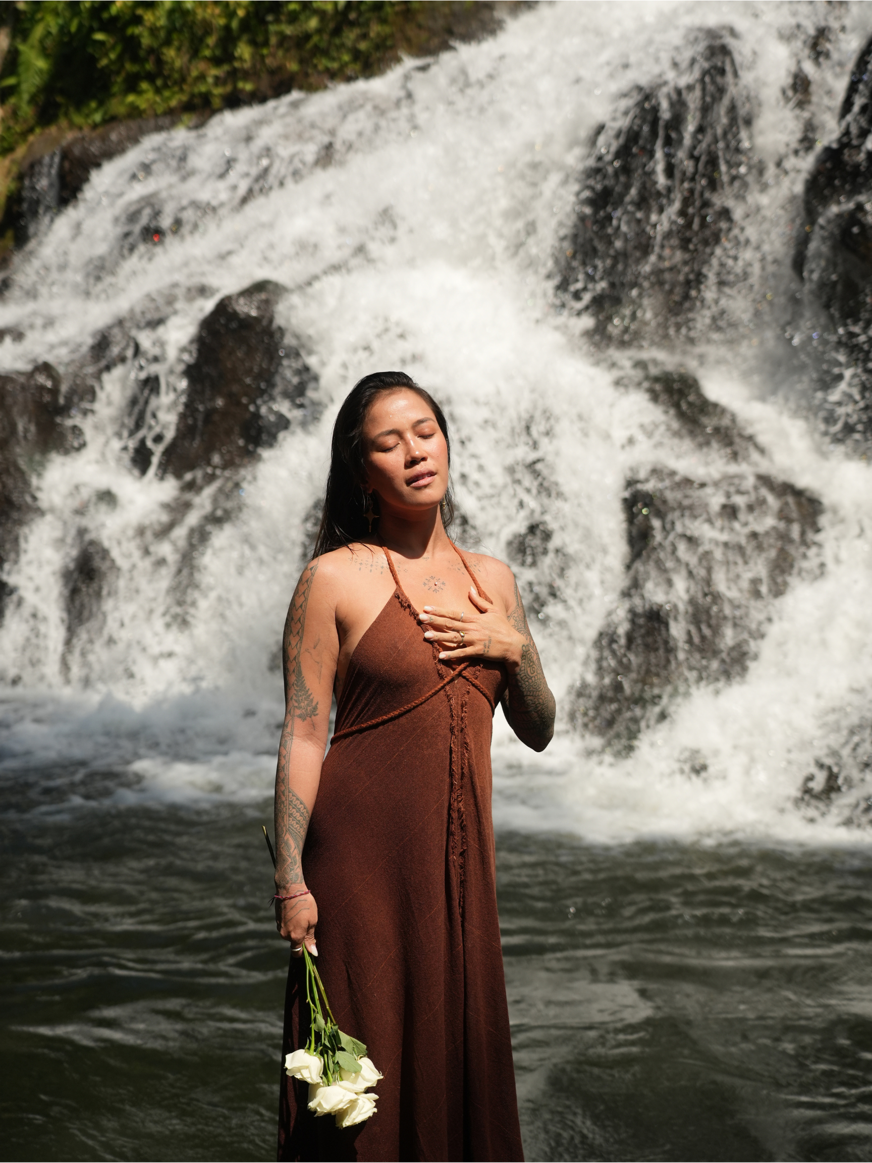 A woman with tattoos on her arms, wearing a brown dress, stands with her eyes closed in front of a waterfall, holding a bouquet of white roses.