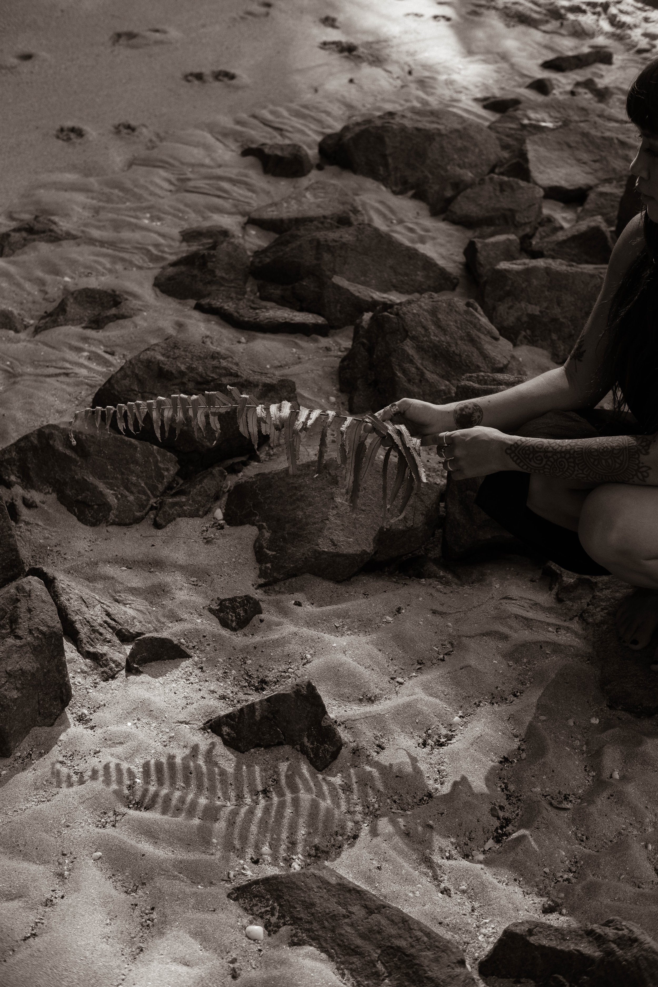 A person with tattoos is sitting on the sandy ground, holding a fish skeleton, with rocks scattered around on the beach.