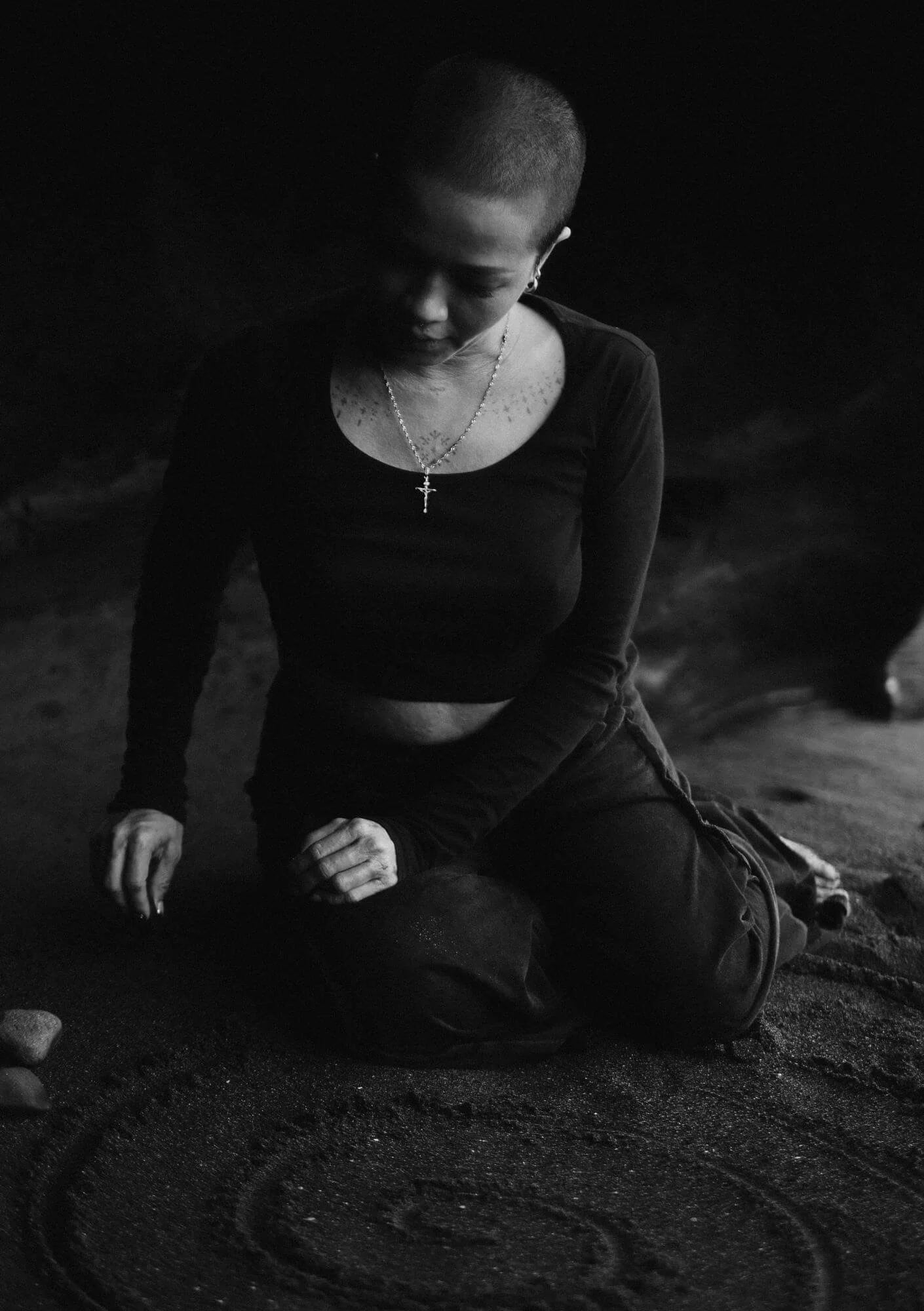 A woman with a shaved head kneels on the sand near the ocean at night, wearing a black long-sleeve top and a necklace with a cross.