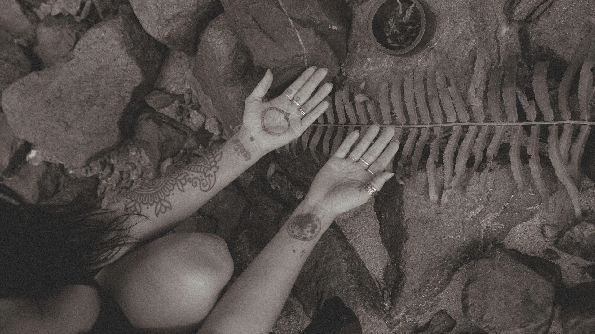 Person with tattoos on arms and hands, displaying circular markings on palms, is lying on rocky ground with plant fossils, possibly fern or palm leaves, nearby.
