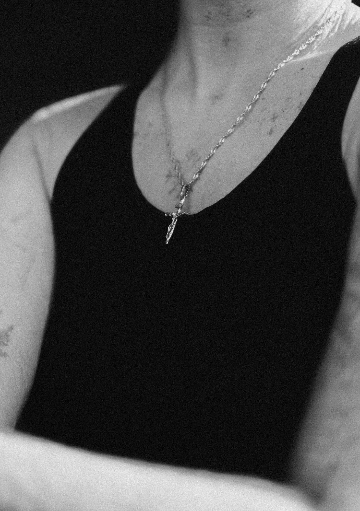 Close-up of a woman wearing a cross necklace and a black sleeveless top, with visible tattoos on her shoulder and chest, in black and white.