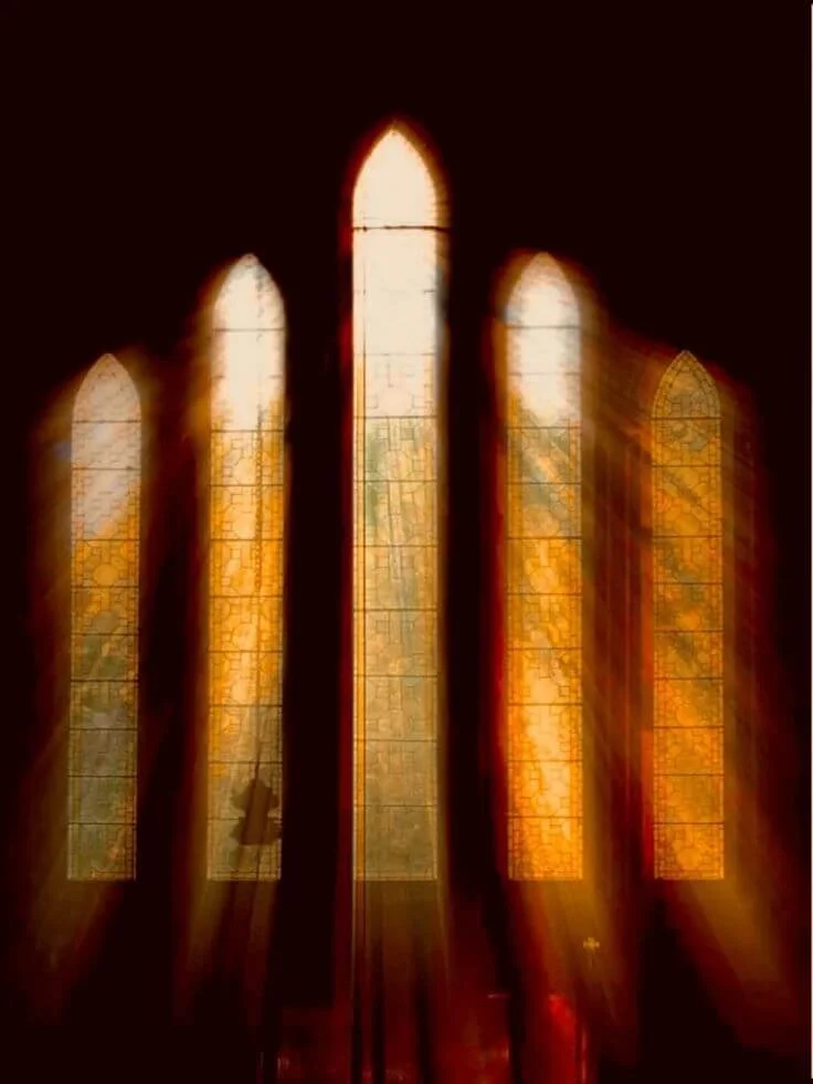 Sunlight shining through five tall stained glass windows with yellow and orange hues in a dark church interior.
