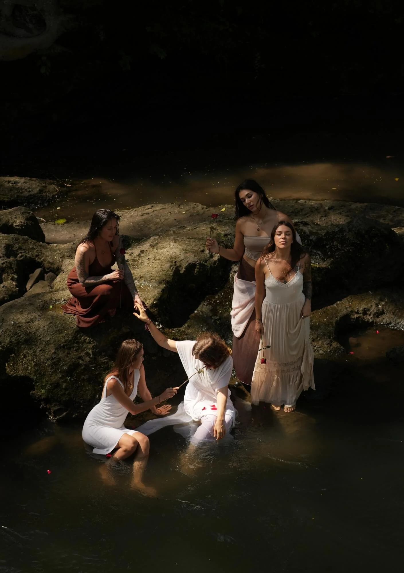 Six women in white and pastel dresses in a dark, rocky river scene, some holding roses and interacting with each other.