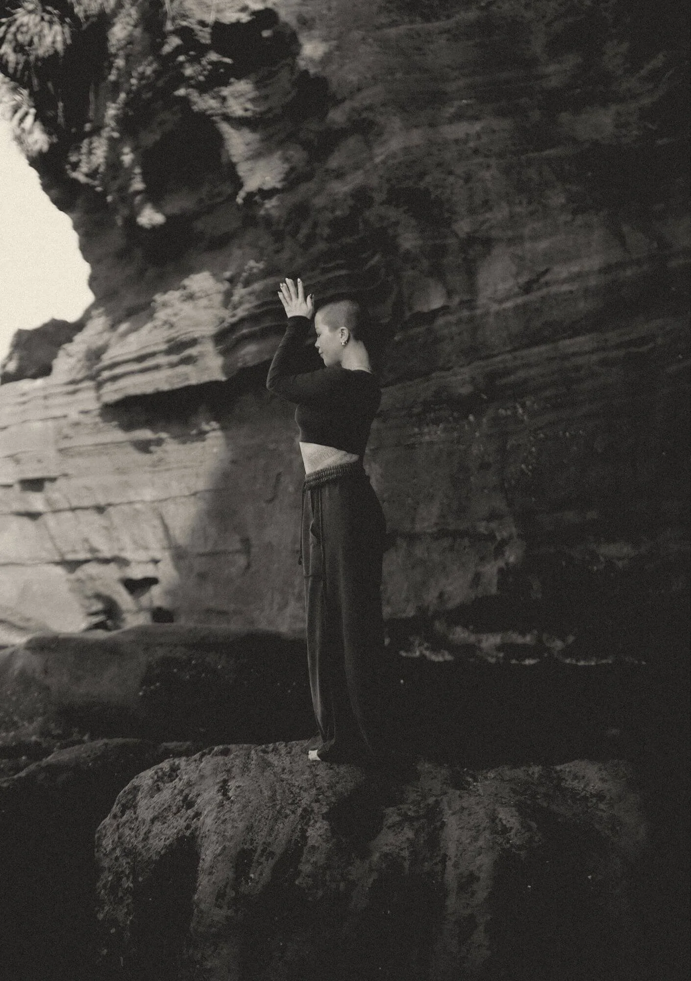 A woman with short hair, dressed in dark clothing, standing on a large rock near a rugged cliff face, with her hands raised above her head, engaging in a pose or meditation near a natural rocky landscape.