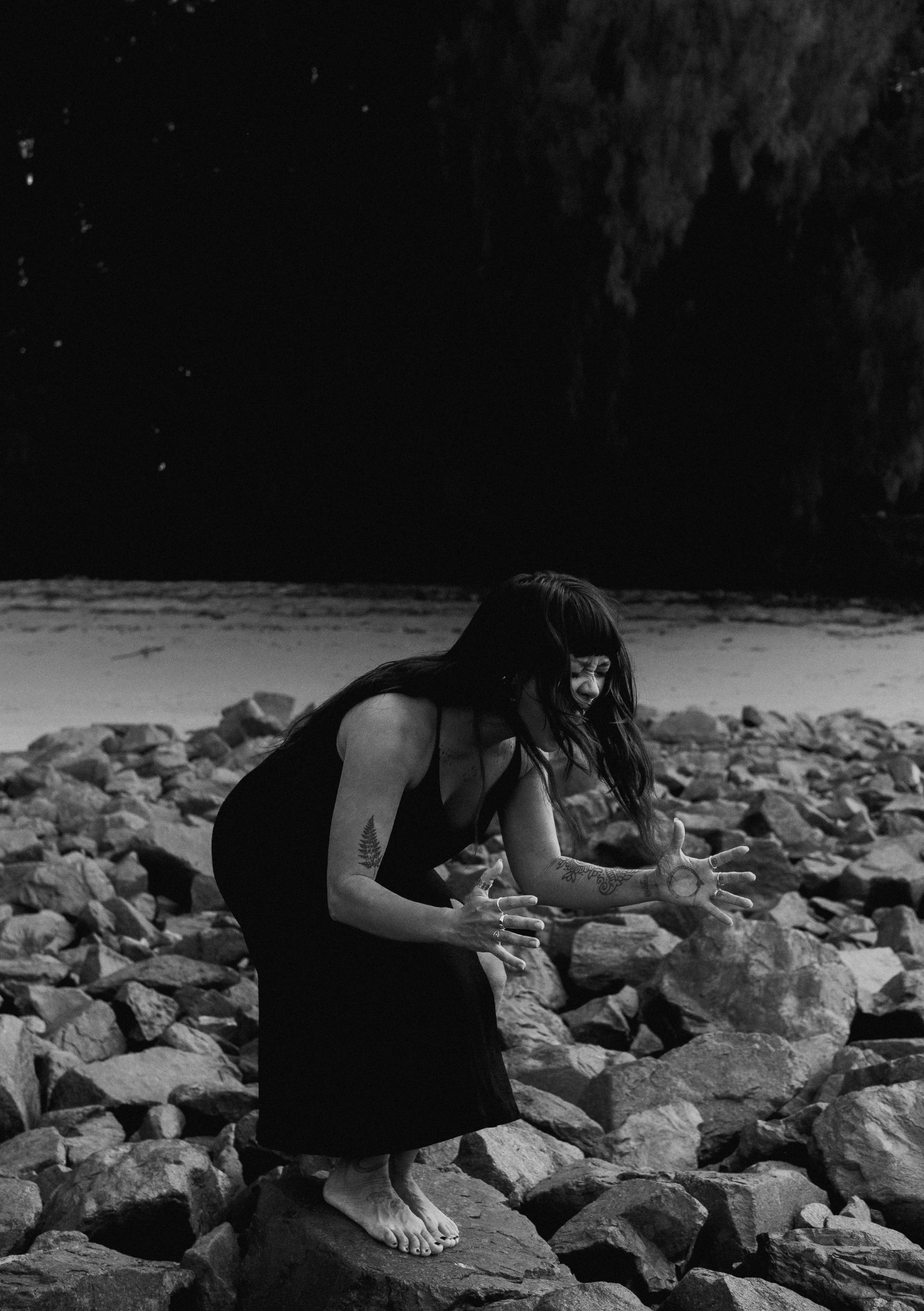 A woman with long dark hair, tattoos on her arms, wearing a black dress, standing barefoot on rocks at night, with a night sky and stars in the background, and trees in the distance.