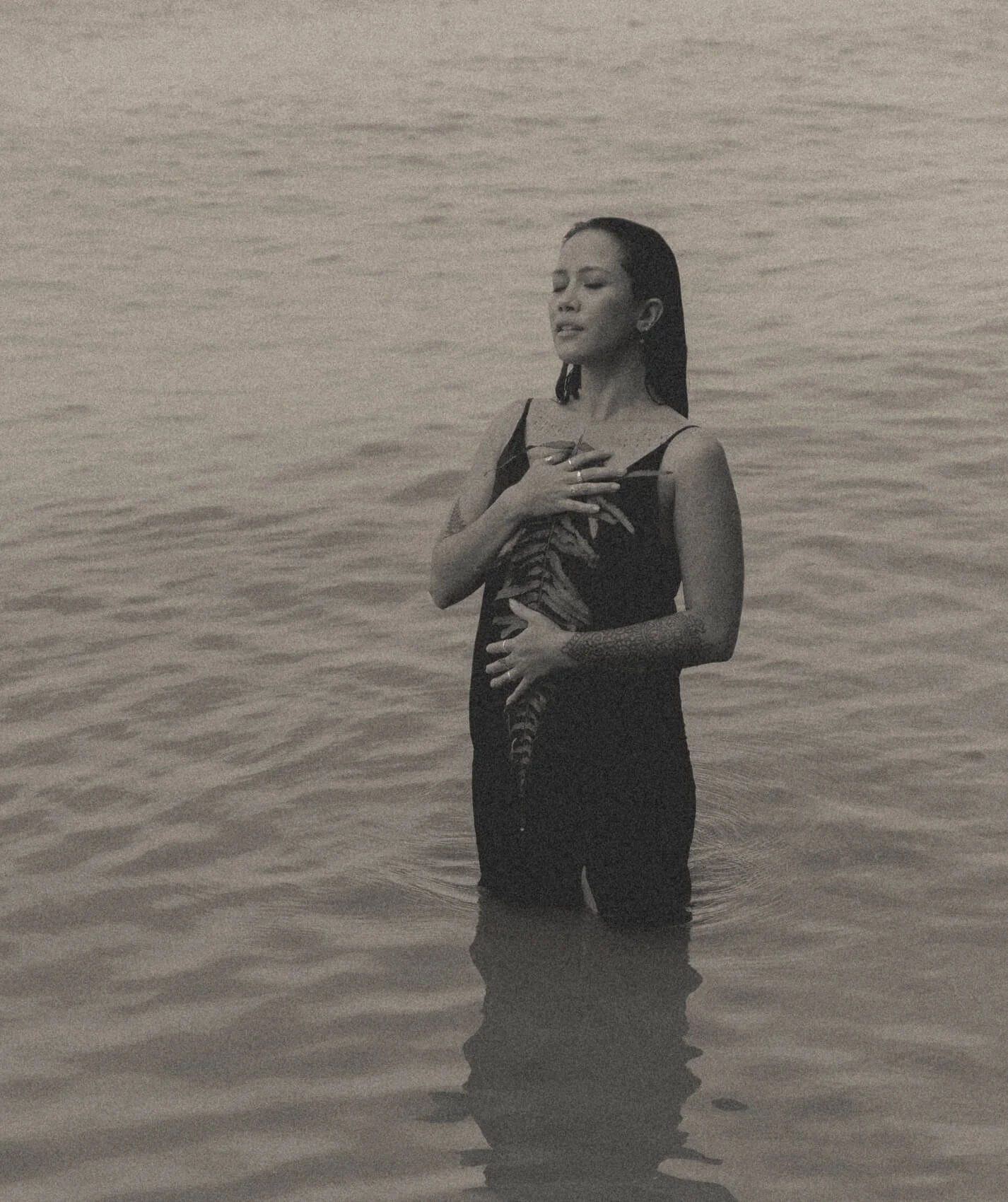 A woman standing waist-deep in water, holding a cat, with her hand on her chest and her eyes closed.