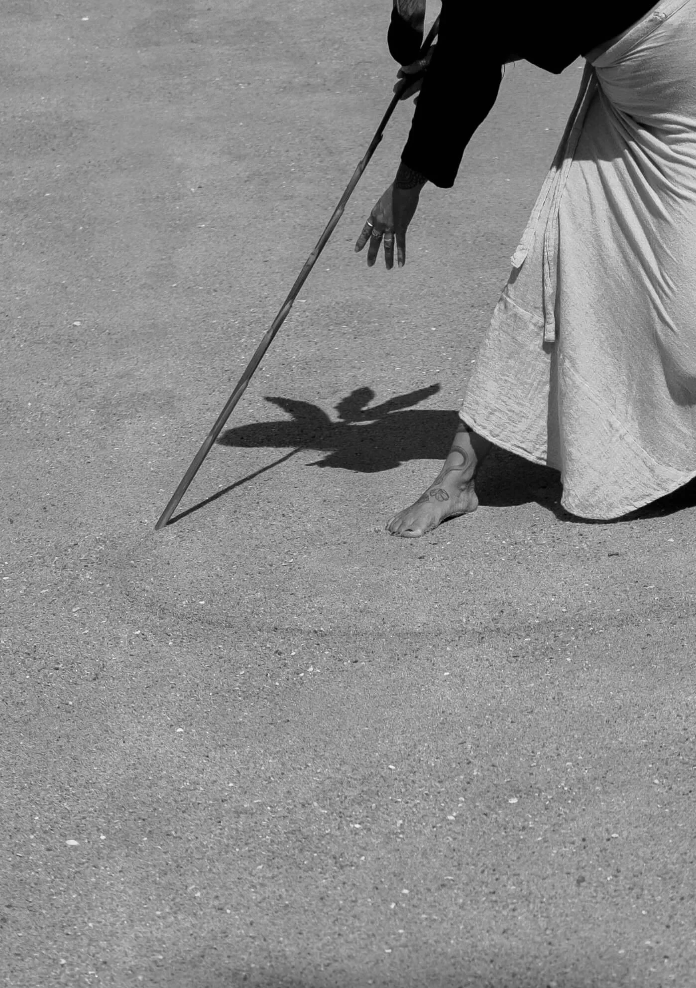 A person with a tattoo on their foot and another on their hand, barefoot, wearing a tie-dye skirt and a black long-sleeve shirt, holding a long stick, casting a shadow of a bird on the ground.