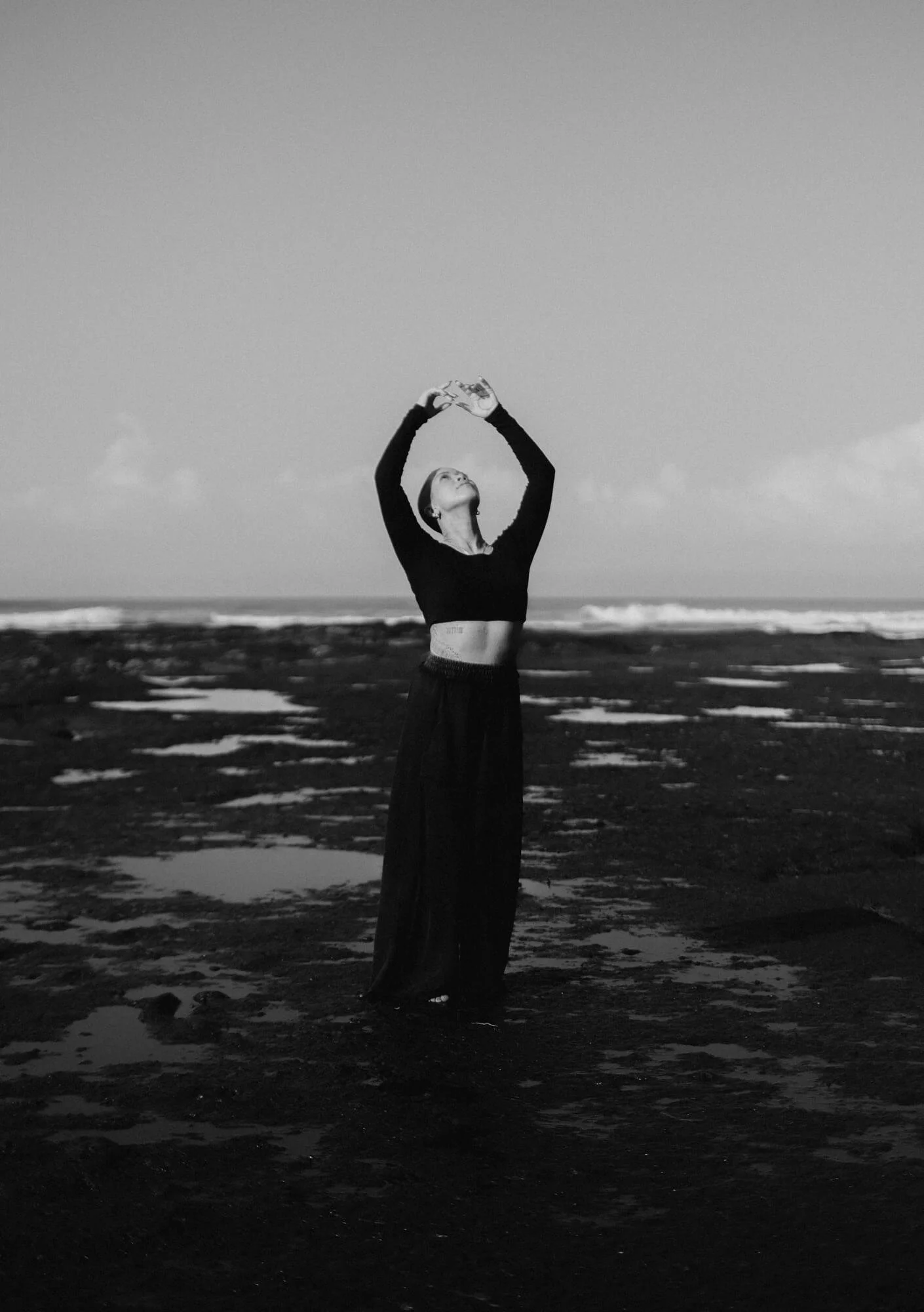 A woman standing on a dark beach holding a camera above her head, looking upwards, with an ocean and cloudy sky in the background.