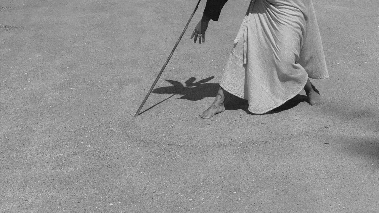 A person in a long skirt kneels on a sandy surface, holding a stick that casts a shadow resembling a bird.