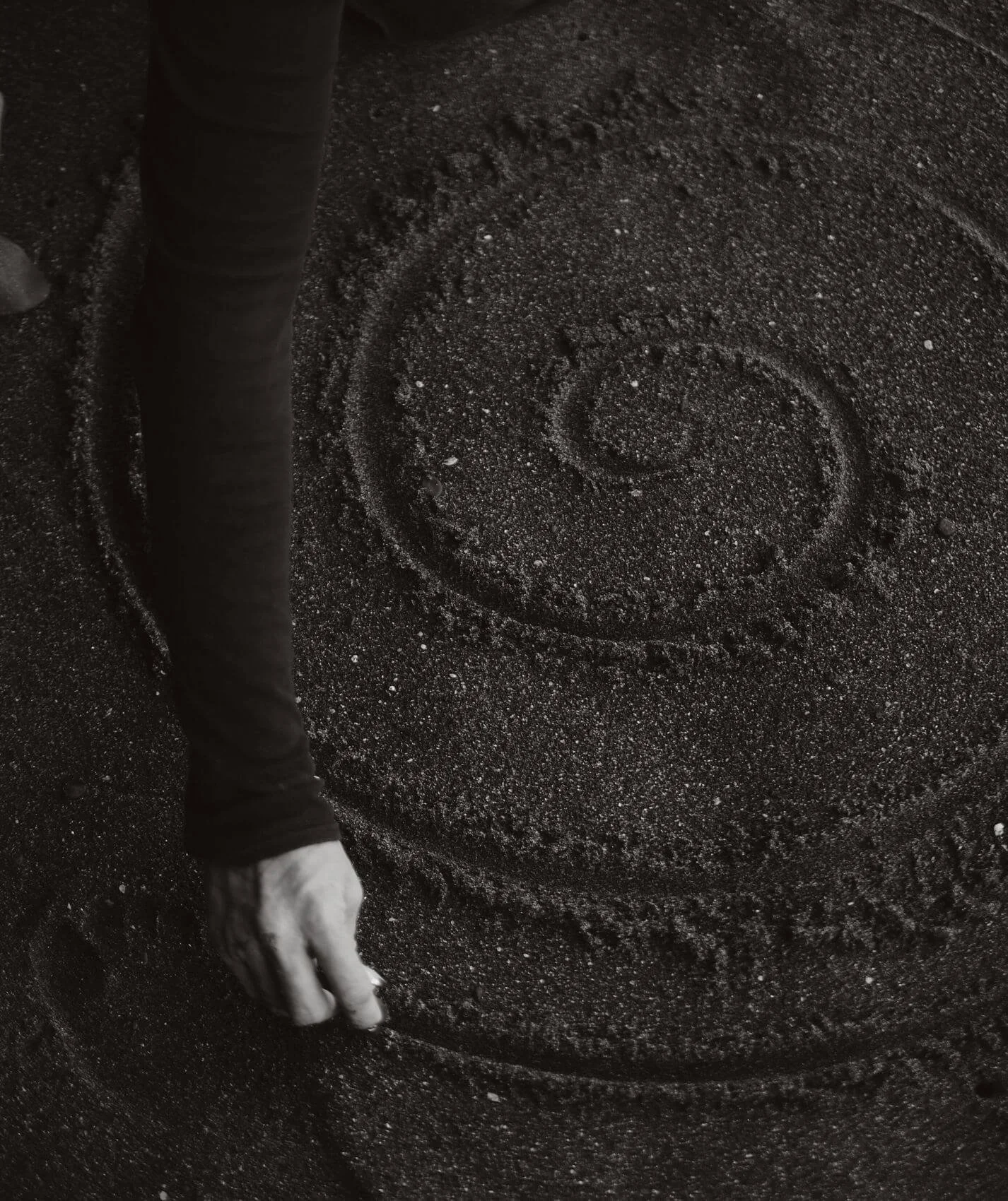 A person's hand is working with black sand to create a spiral design while wearing a long-sleeved shirt.