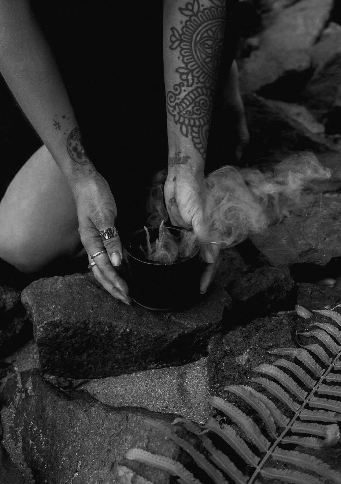 A person with tattooed arms holding a steaming cup placed on rocks outdoors, with a fern plant visible in the foreground.