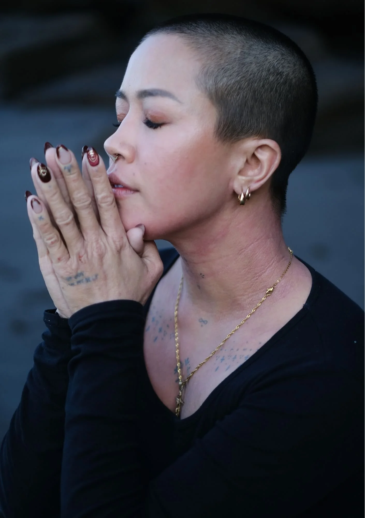 A woman with a shaved head praying or meditating with her hands clasped near her face, wearing multiple earrings and layered necklaces.