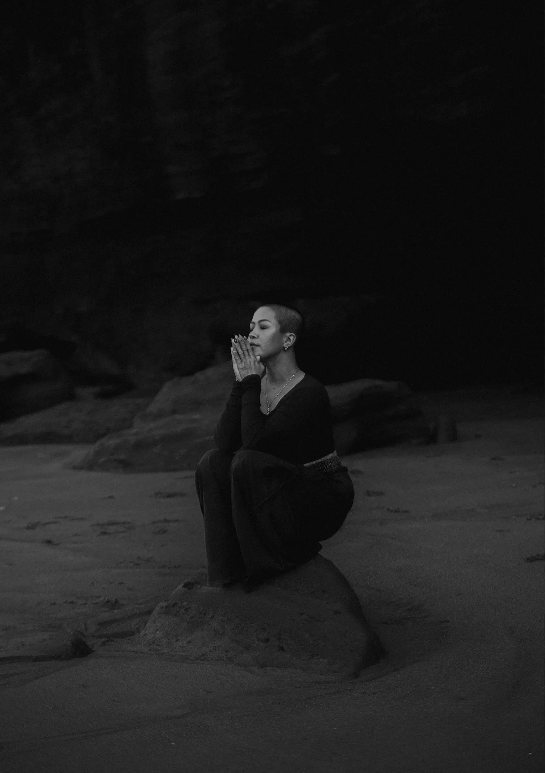 A woman with a shaved head kneeling on a rock at a beach, with her hands pressed together near her face, eyes closed, in a meditative pose, in black and white.