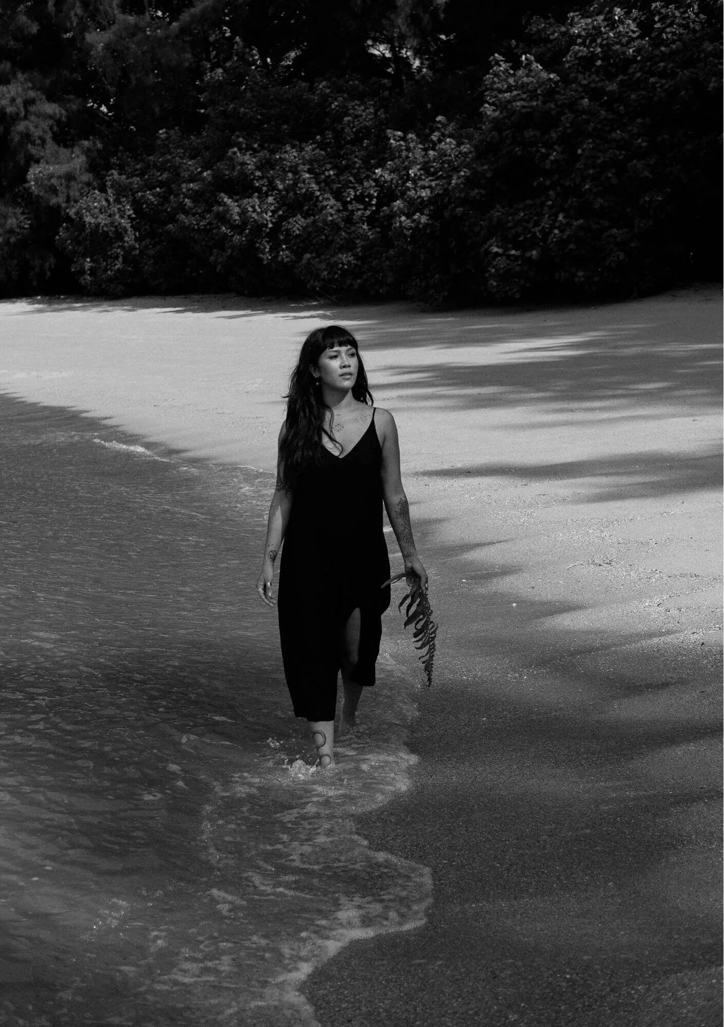A woman in a black dress walking in shallow ocean water along a sandy beach with trees in the background, black and white photo.