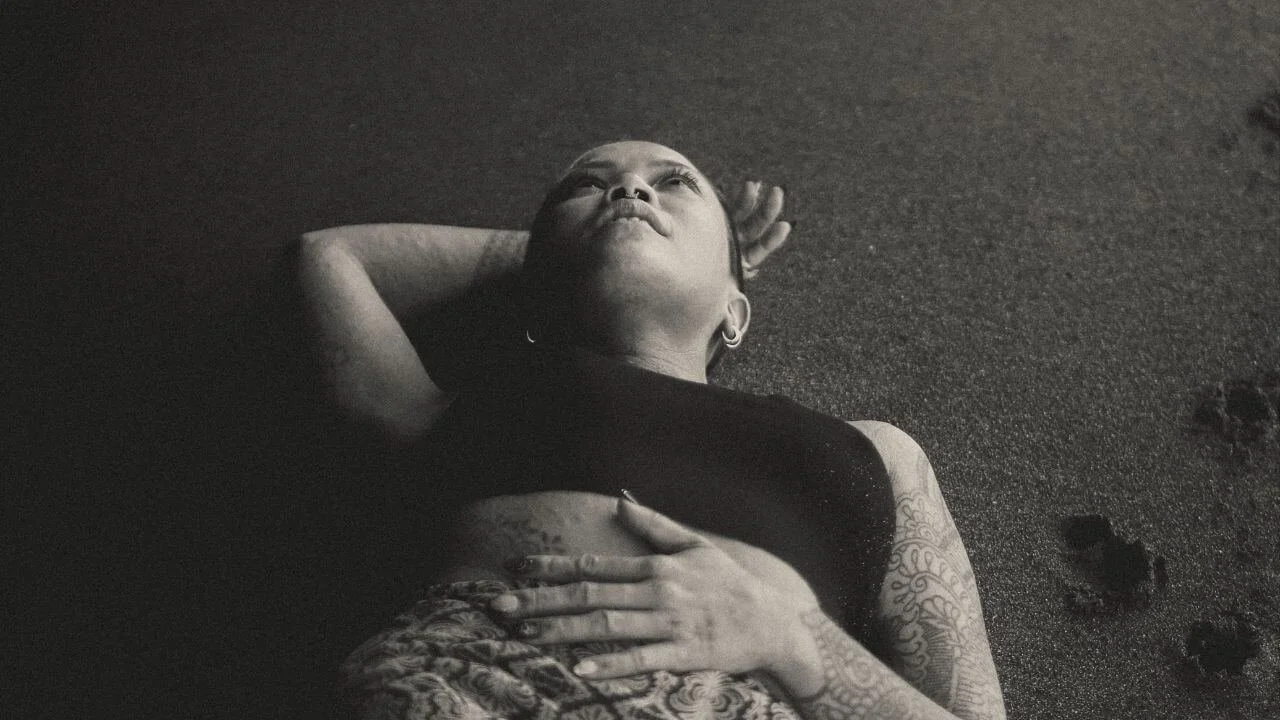 A woman lying on her back on the sand, looking up, with one hand on her chest and the other behind her head, tattoos visible on her arm, wearing earrings and a sleeveless top.