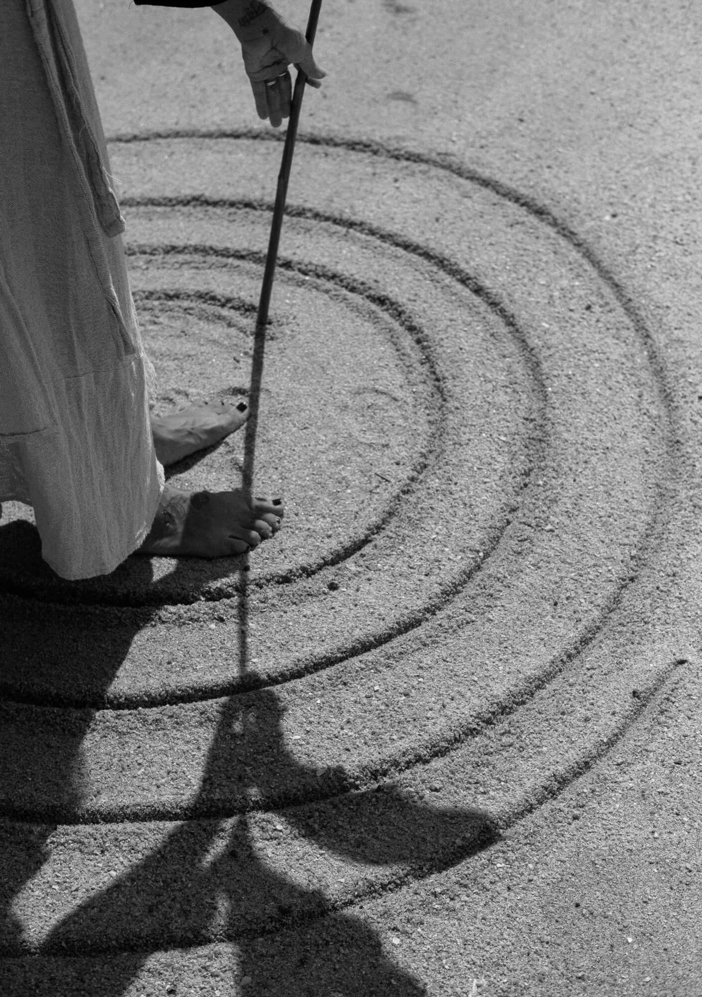 A person standing barefoot on sandy ground, drawing concentric circles with a stick, with a shadow cast on the sand.