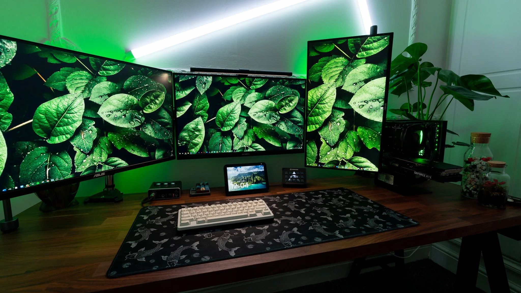 A desk setup with three large monitors displaying green leaf images, a mechanical keyboard, a small digital clock, and various electronic accessories. There are green LED lights behind the monitors and potted plants on the desk and corner of the room.