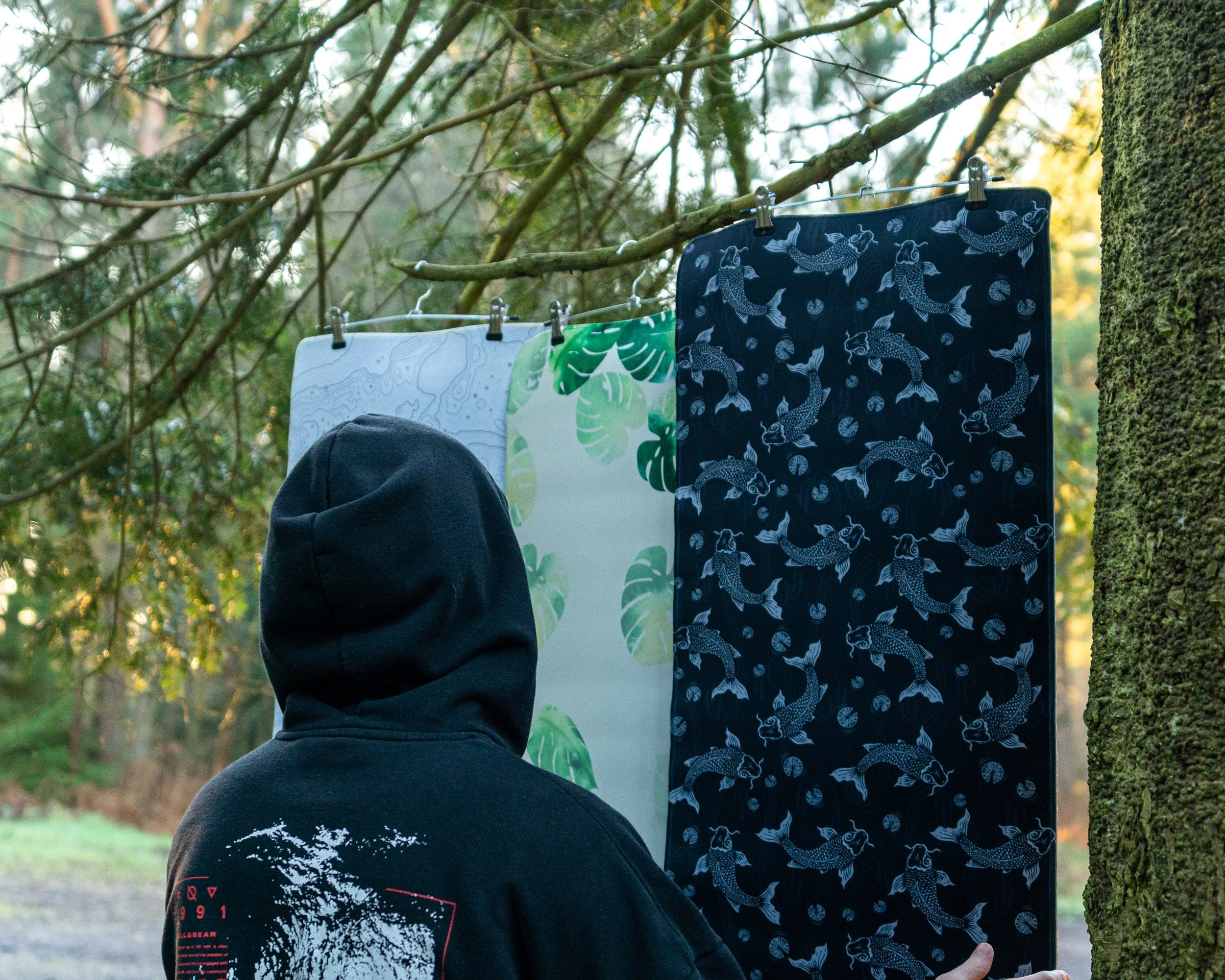 Person wearing a black hoodie with a graphic on the back, standing outdoors near trees, looking at colorful hanging fabric or scarves with patterns of fish and leaves.