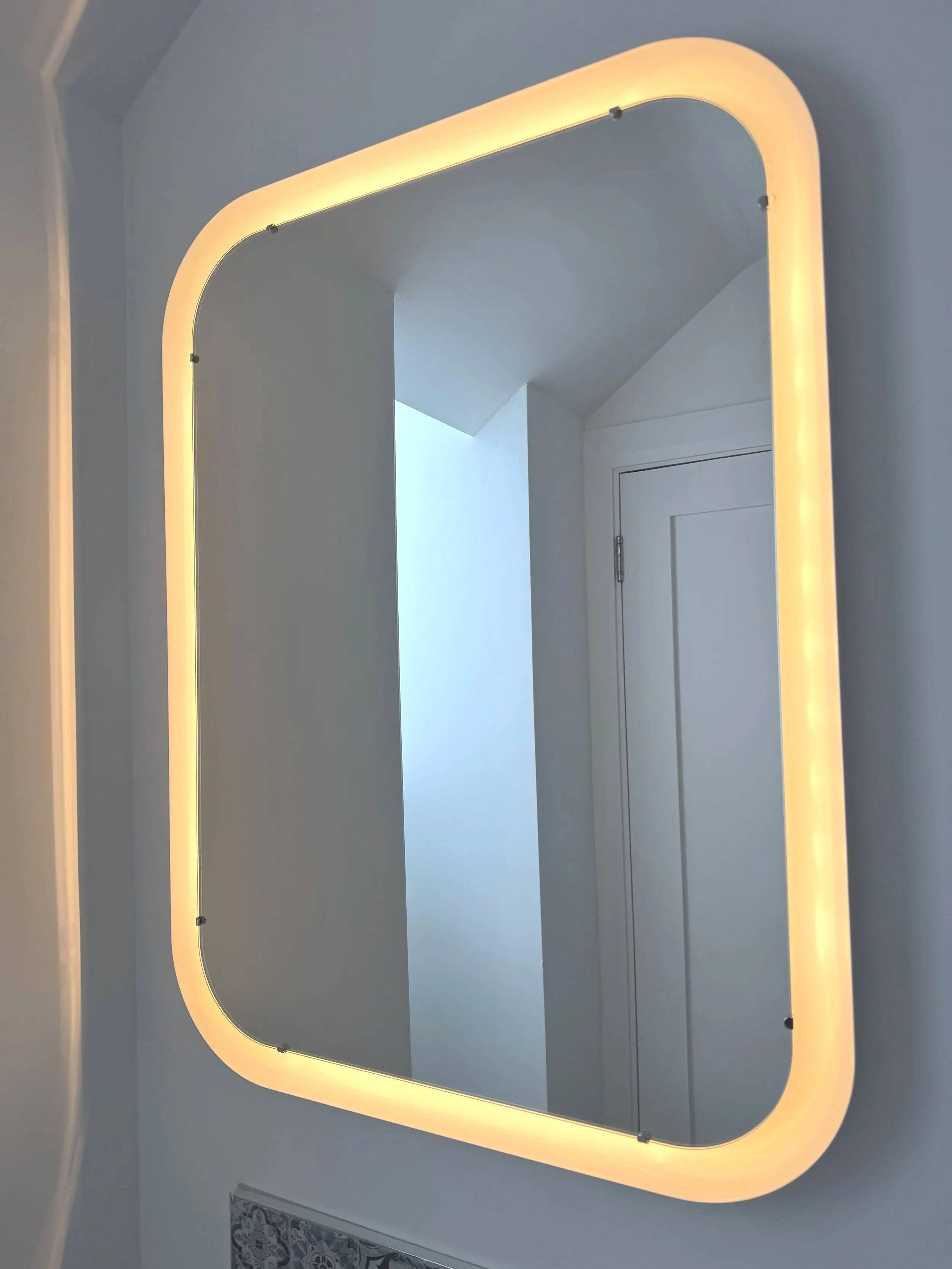 A rectangular mirror with rounded corners and an integrated LED light frame mounted on a wall above a section with patterned tile.