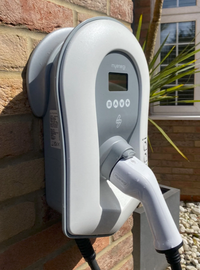 Electric vehicle charging station mounted on a brick wall outdoors with a potted plant and window in the background.