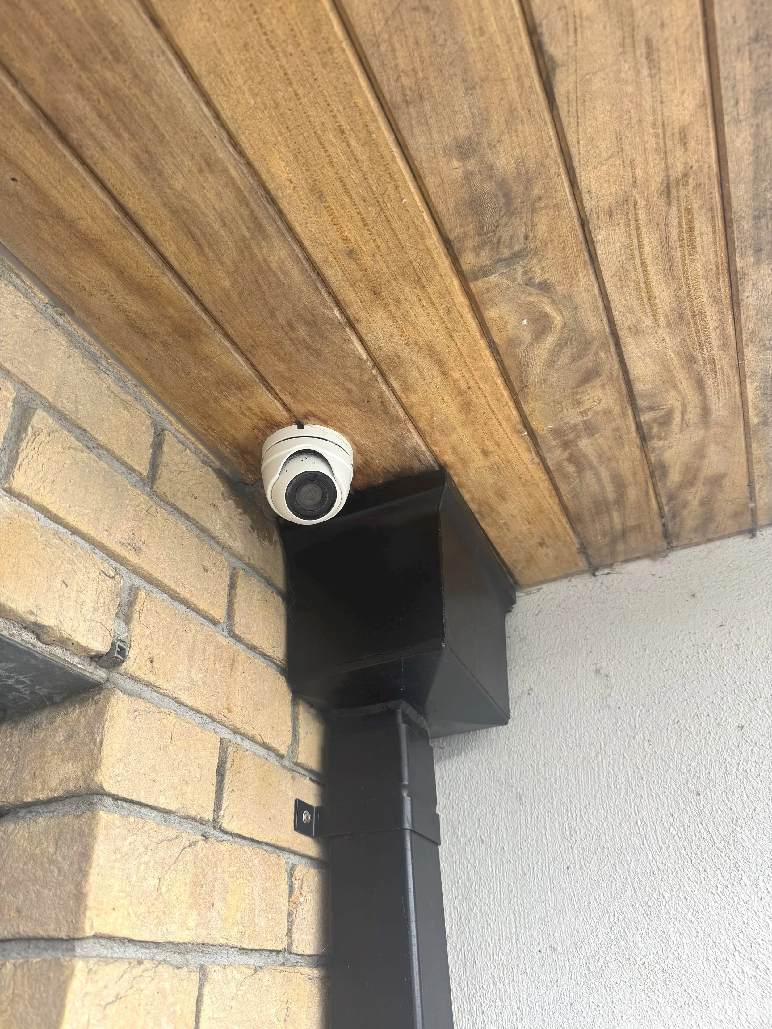 Security camera mounted on brick wall below a wooden ceiling, near a black vent pipe.