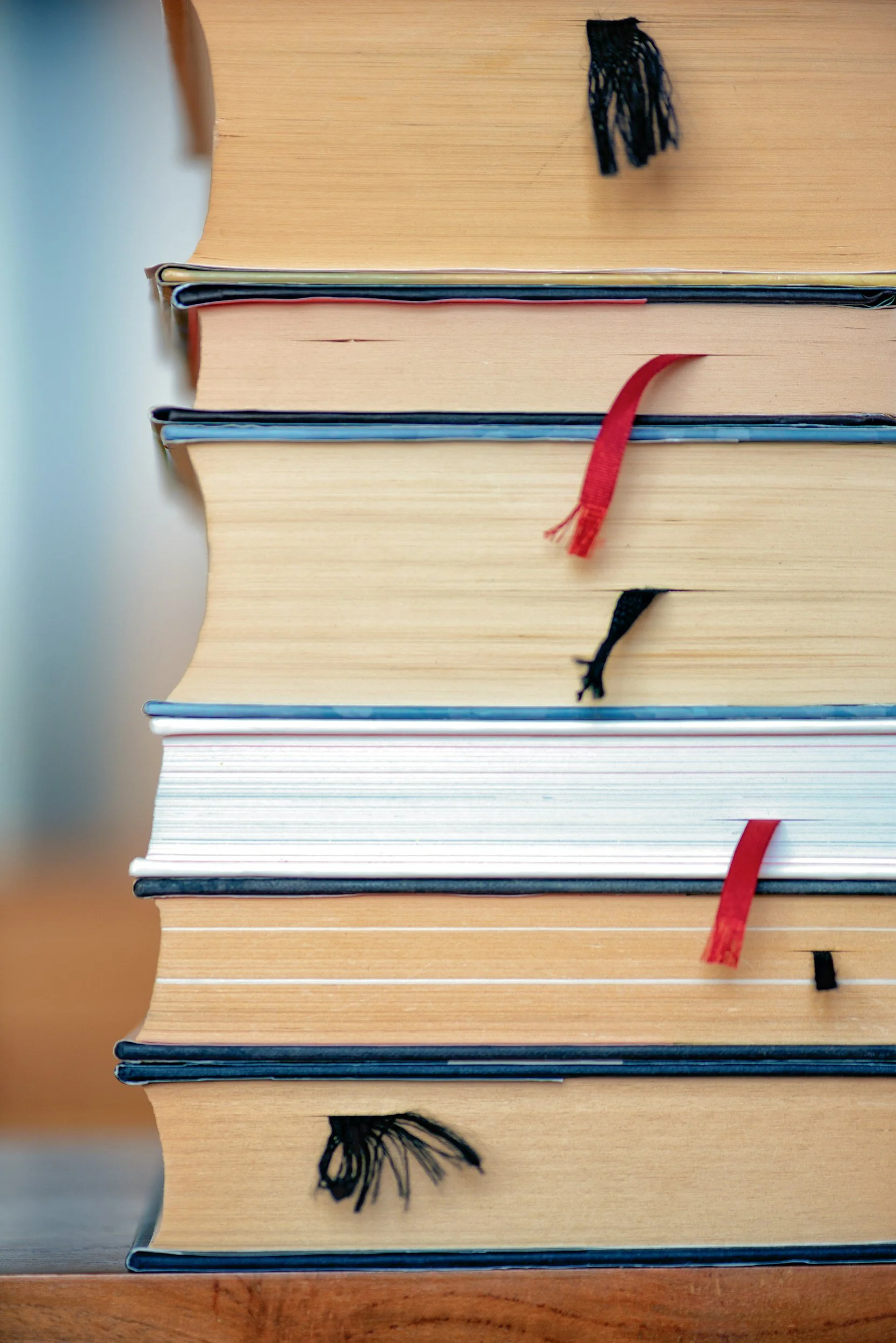 A stack of seven thick hardcover books on a wooden surface, with various colored bookmark ribbons protruding from the pages.
