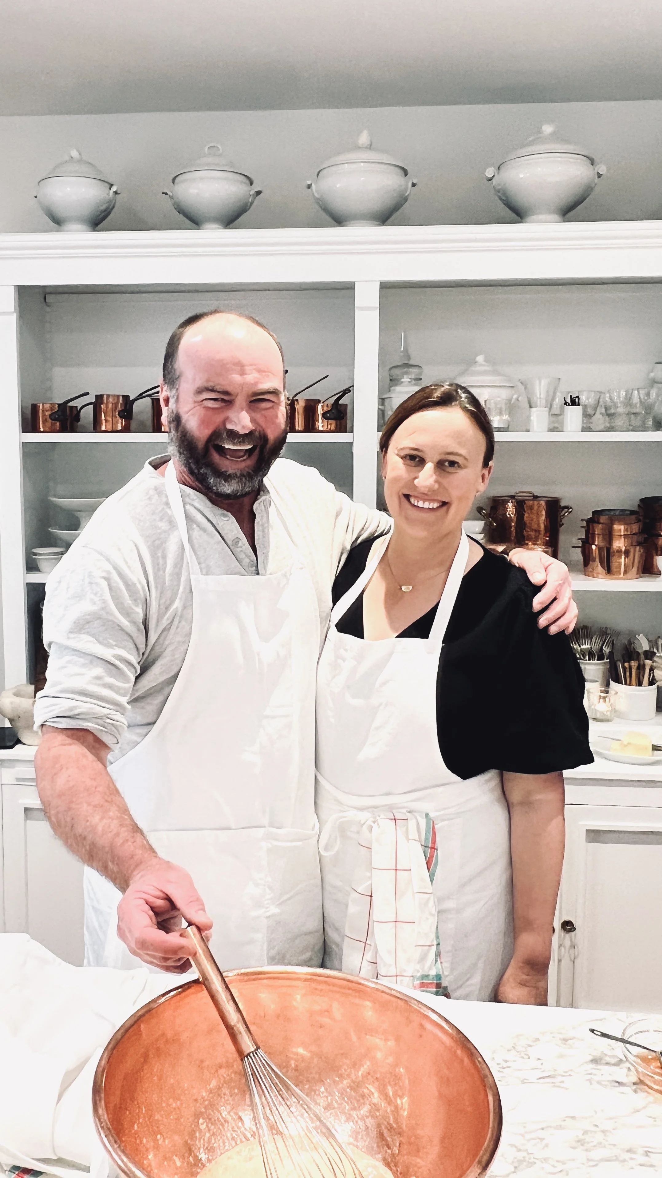 Courtney and Con Watts wearing aprons in a French kitchen, smiling and posing for a photo while cooking together.