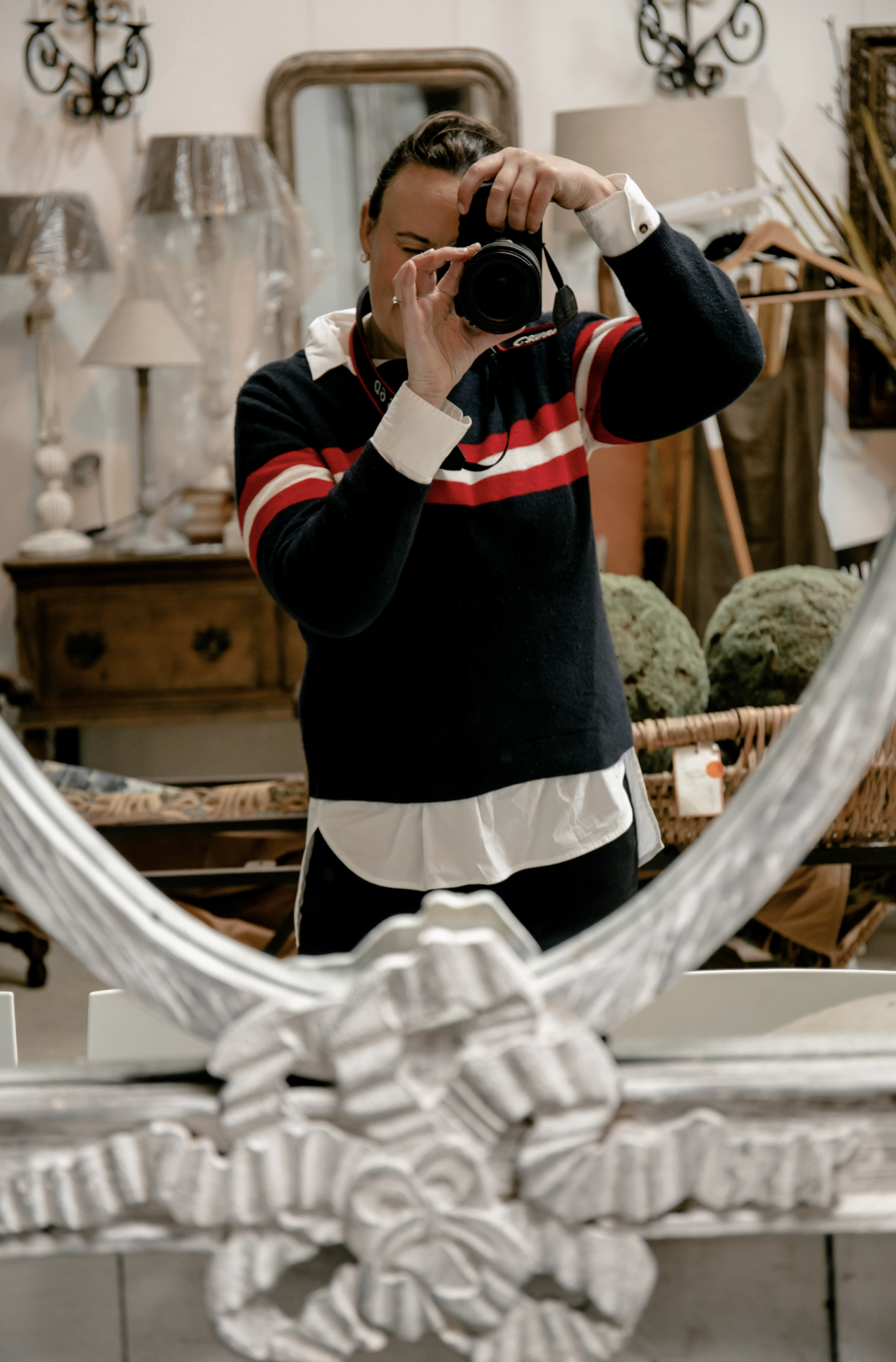 Courtney Watts taking a photo with her camera of a decorative silver object or sculpture, reflected in a mirror, with a background of lamps, framed pictures, and furniture in a room.