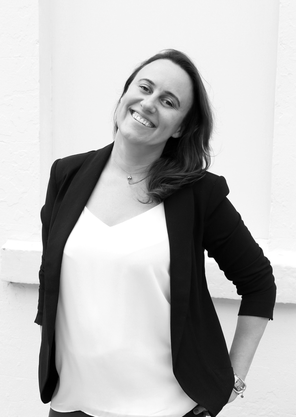 Black and white portrait of Courtney Watts Luxury Travel Concierge smiling, wearing a black blazer over a white top, standing in front of a plain white wall.