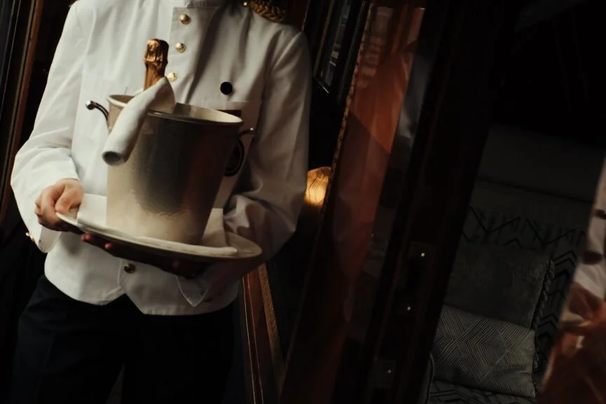 Man in a crisp white uniform holding a tray with a metal ice bucket and a bottle of French champagne. He is on a Belmond Train.