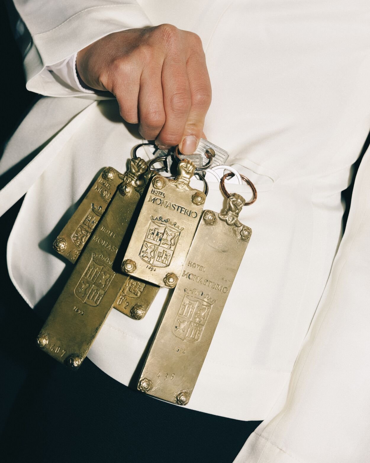 A person's hand holding several vintage-style keychains shaped like hotel name tags with the name 'Hotel Monasterio' and a coat of arms, attached to rings.