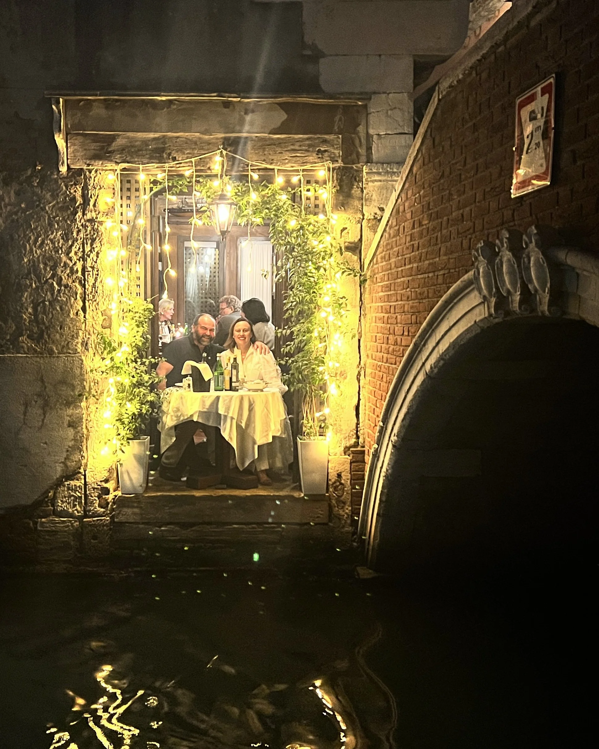 Courtney and Con Watts dining at a table inside a cozy, dimly lit restaurant or bar, seen through a window decorated with string lights. Their table overlooks the canal in Venice.