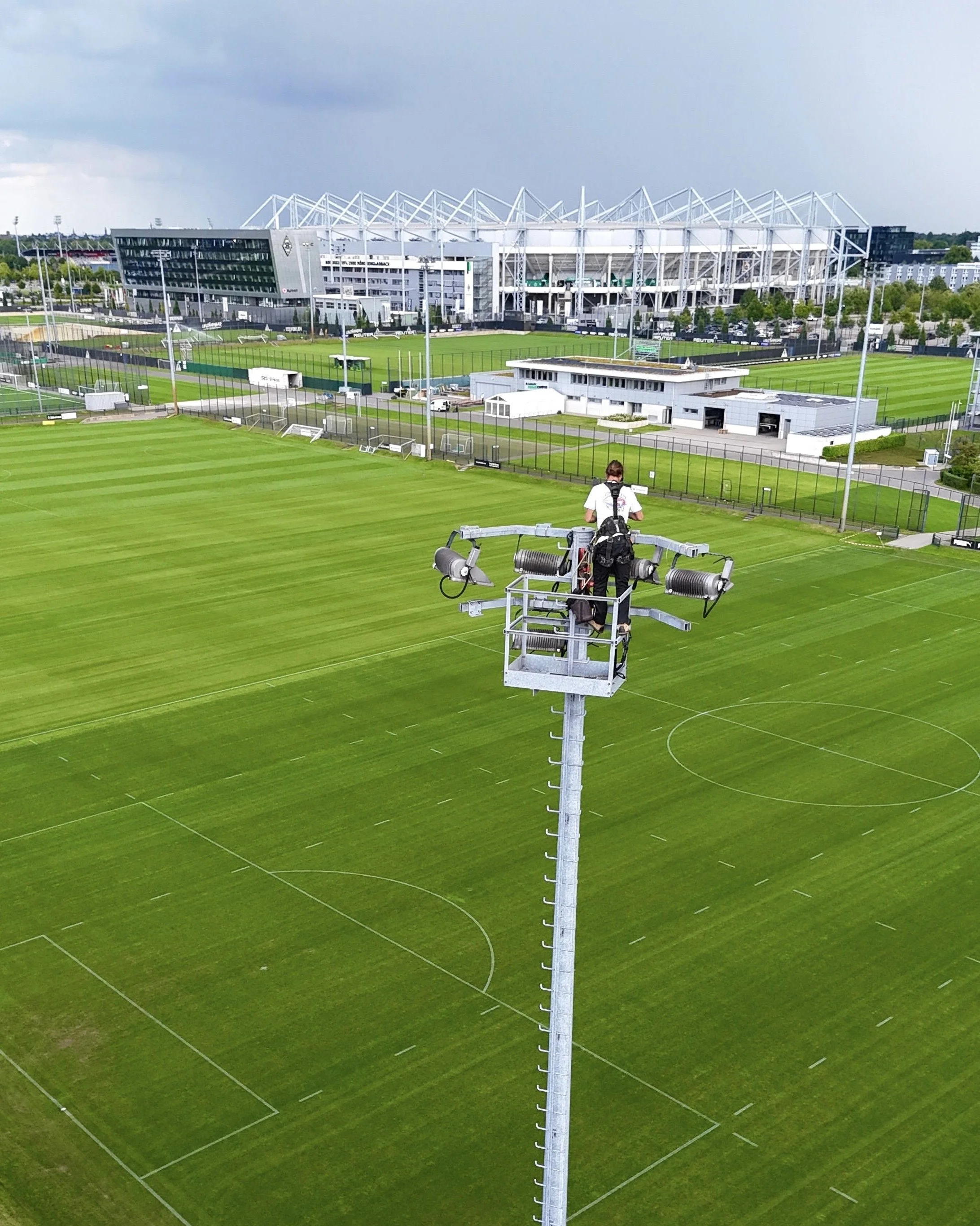 A person in tactical gear standing on a tall stadium light pole, inspecting or maintaining the floodlights on a soccer field with freshly mowed grass. In the background, a large modern sports stadium and several buildings are visible.