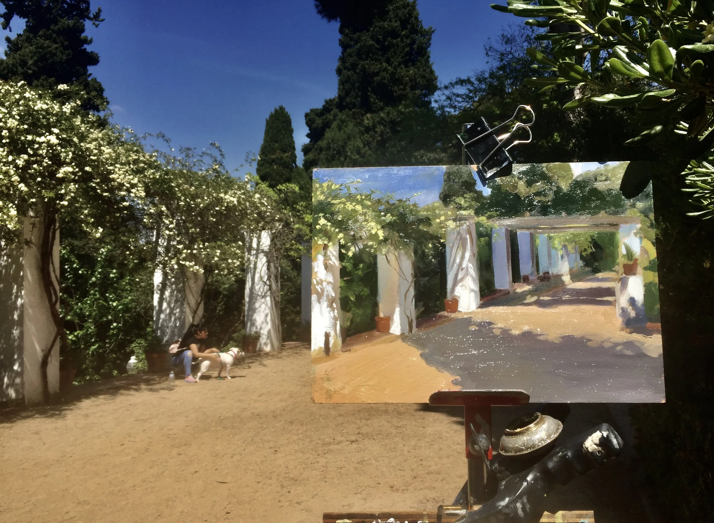 Outdoor scene of a person sitting on a bench with a dog in a garden, with a painting of the same scene on an easel in front of them. The garden has white trellises, green foliage, and tall trees under a bright blue sky.