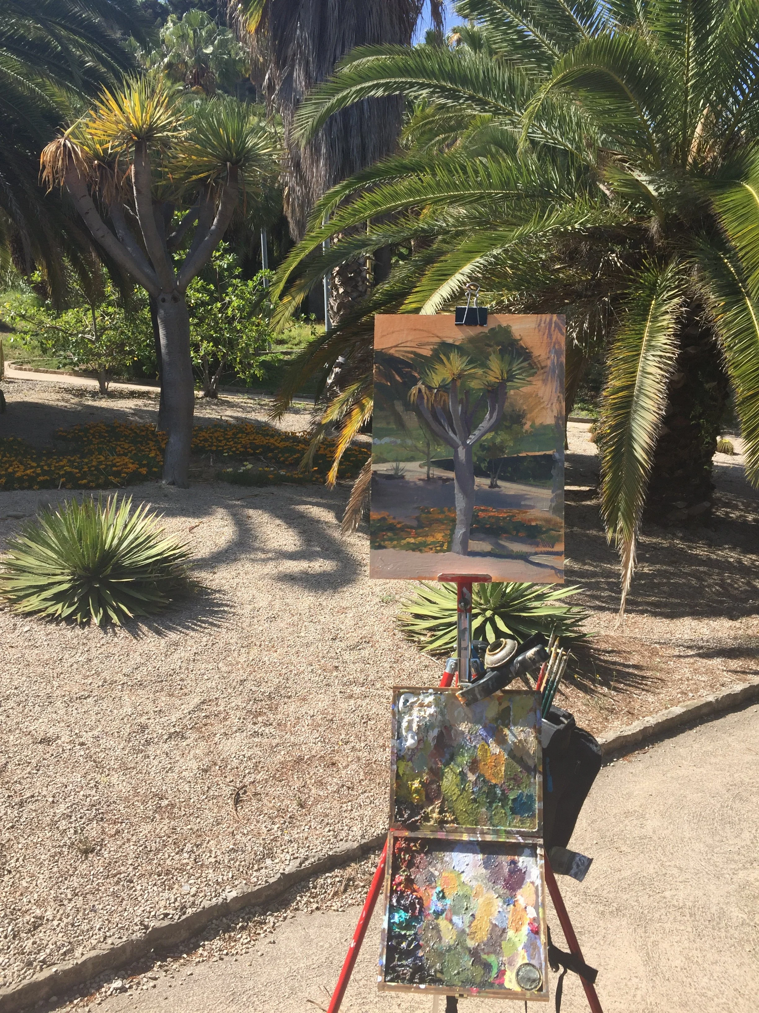 An outdoor artist's easel with a painting of a tree is set up in a sunny park. The painting depicts a tree with a thick trunk and green foliage, similar to the real trees in the background. The easel has a palette with various colors of paint on it, 