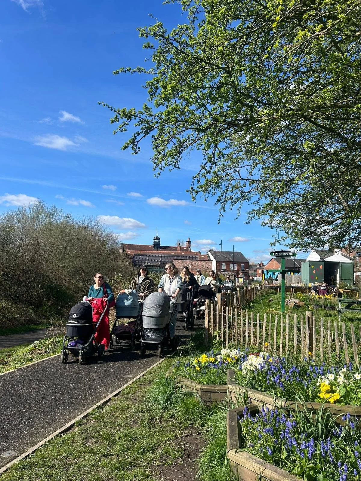 Mums and babies on a walk in Levenshulme on maternity leave. Blue skies and a community garden.