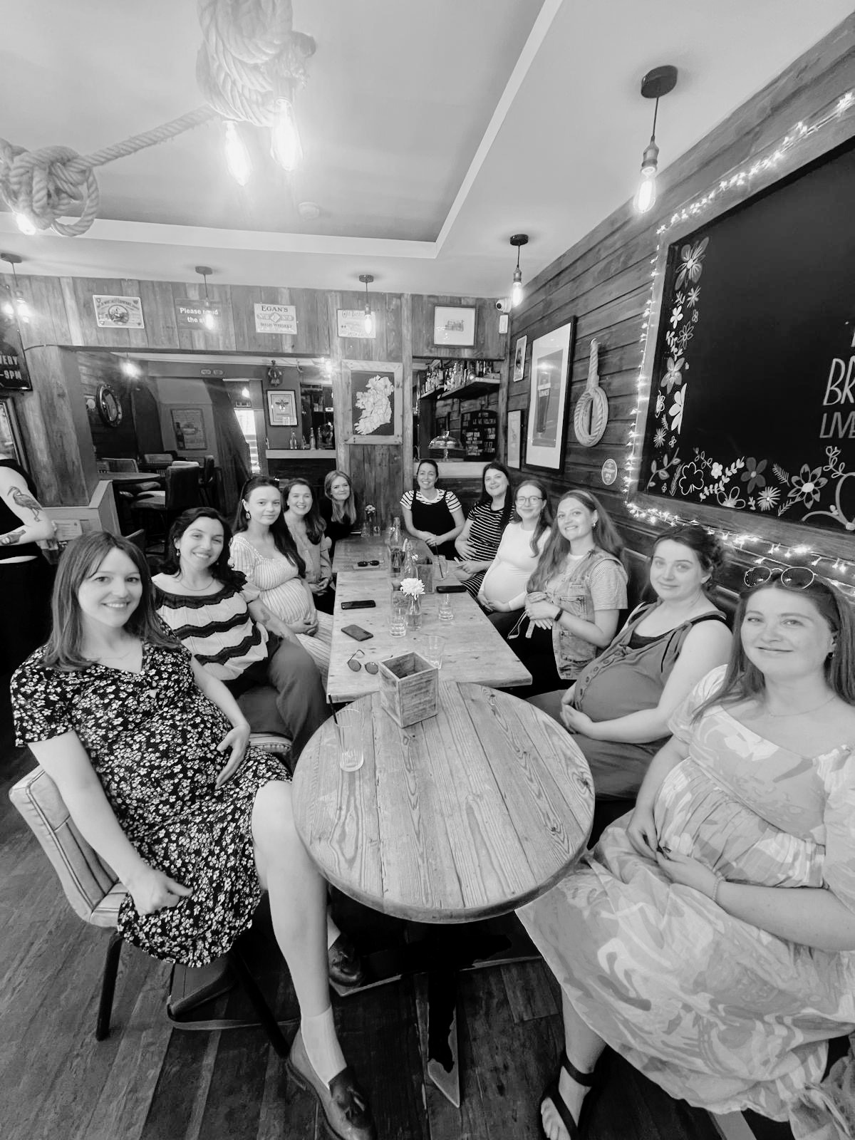 A group of women sitting around a long wooden table in a cozy restaurant with wood-paneled walls, framed pictures, and hanging lights at an antenatal class social event.
