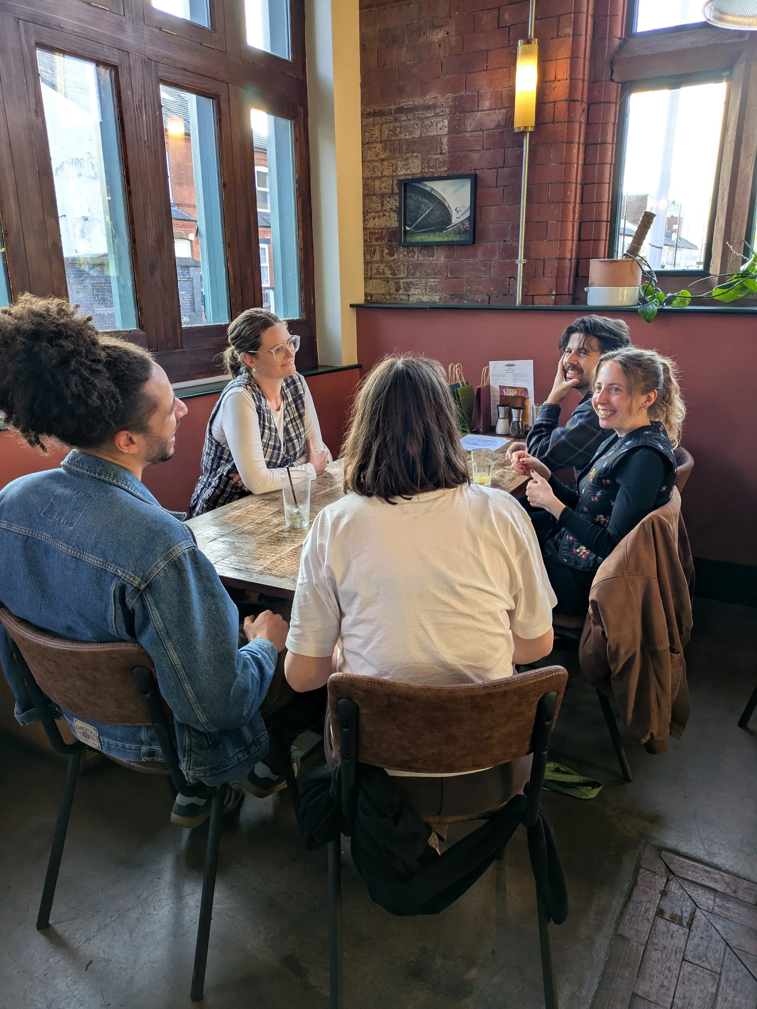 A group of five people sitting around a wooden table having a conversation in a cozy cafe with exposed brick walls, large windows, and natural daylight.