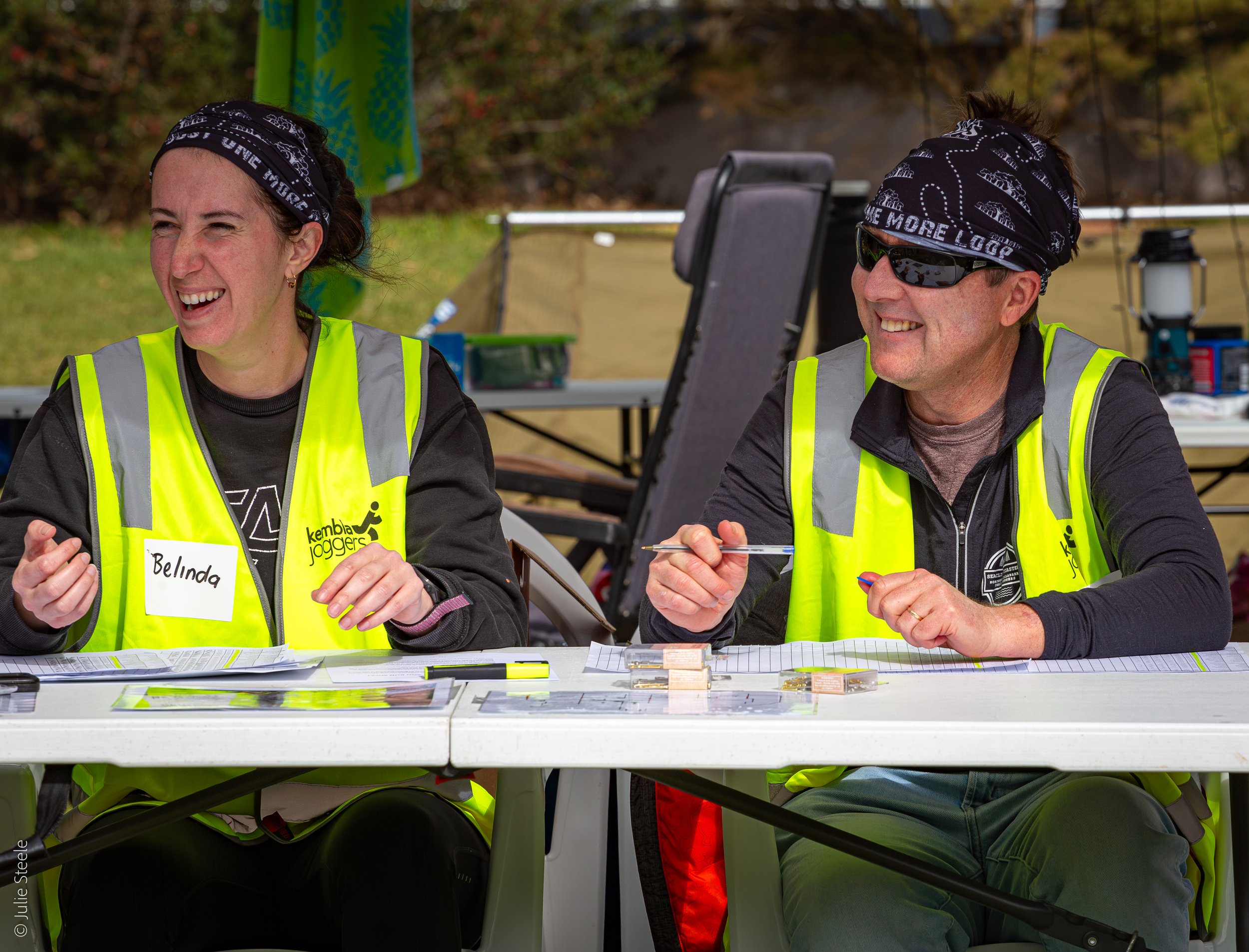 Two adults sitting at a registration table outdoors, both wearing high-visibility vests and black headbands, with one woman smiling and the man wearing sunglasses, indicating they are registering or managing an event.
