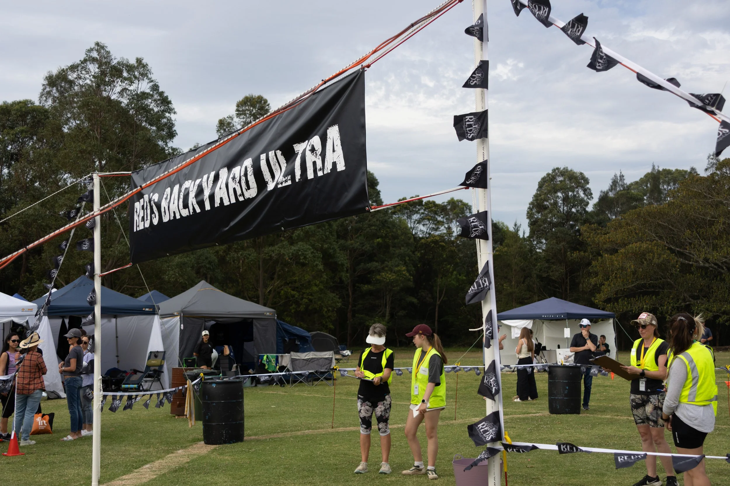 People gathered at an outdoor event, with tents set up in the background. A large black banner reads 'REVS BACKYARD ULTRA,' decorated with smaller flags along the top and side.
