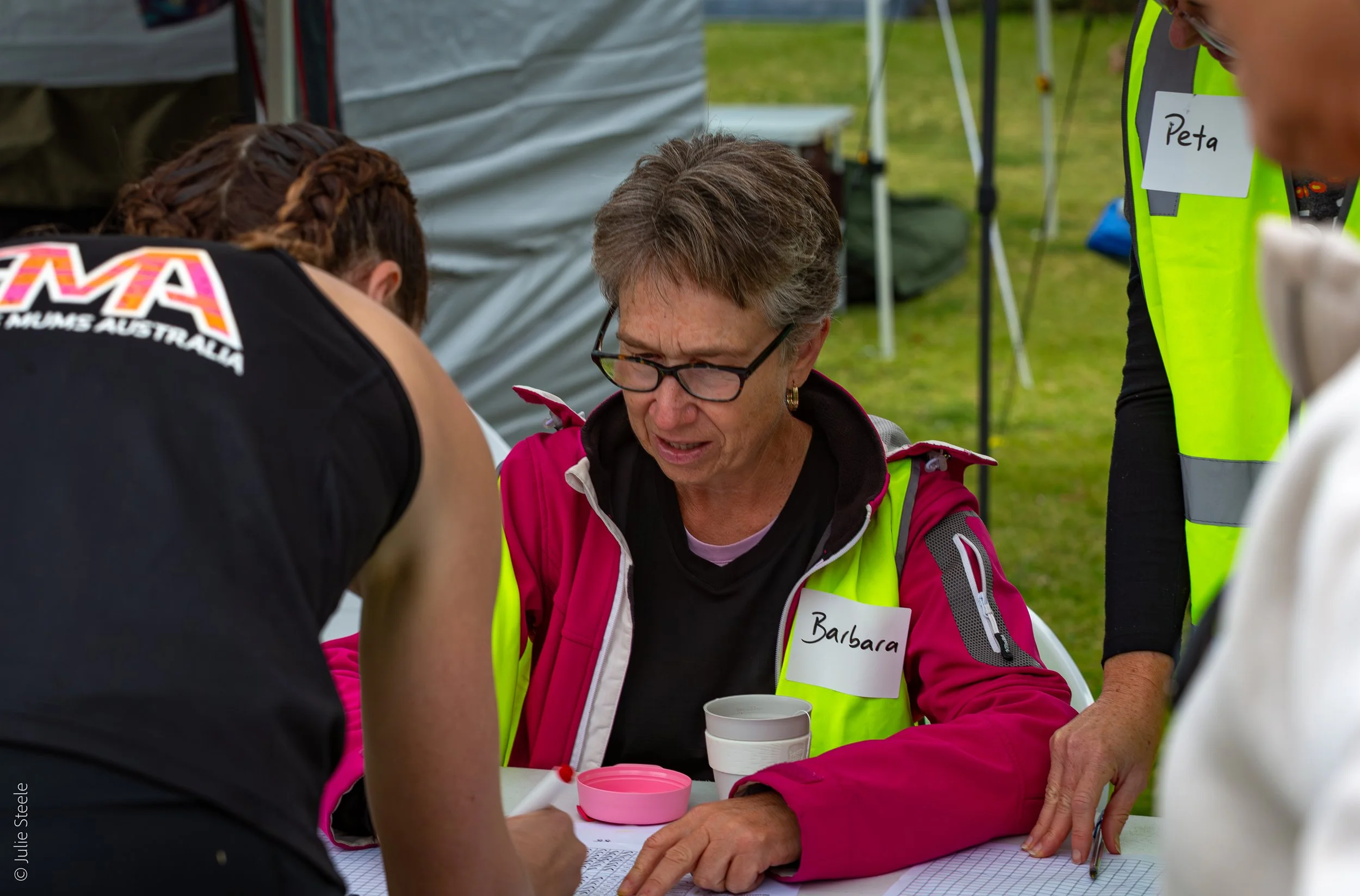 A woman named Barbara, wearing a bright pink jacket and glasses, is sitting at a table with a cup and notes, looking concerned or focused as she interacts with another person during an outdoor event.
