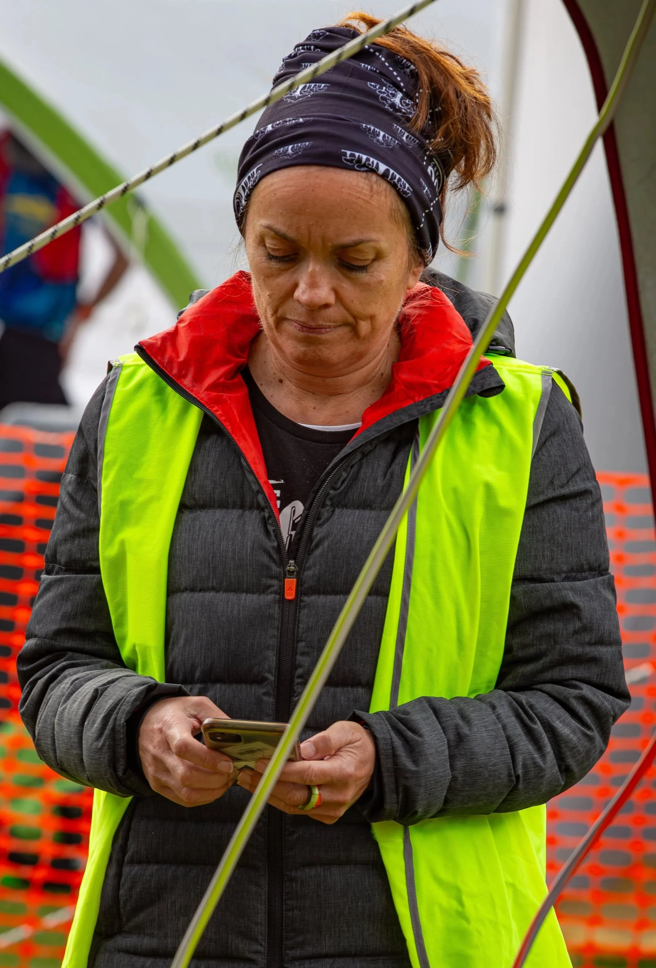 A woman wearing a black jacket with red lining, a neon yellow safety vest, and a black bandana on her head, looking at her phone outdoors.