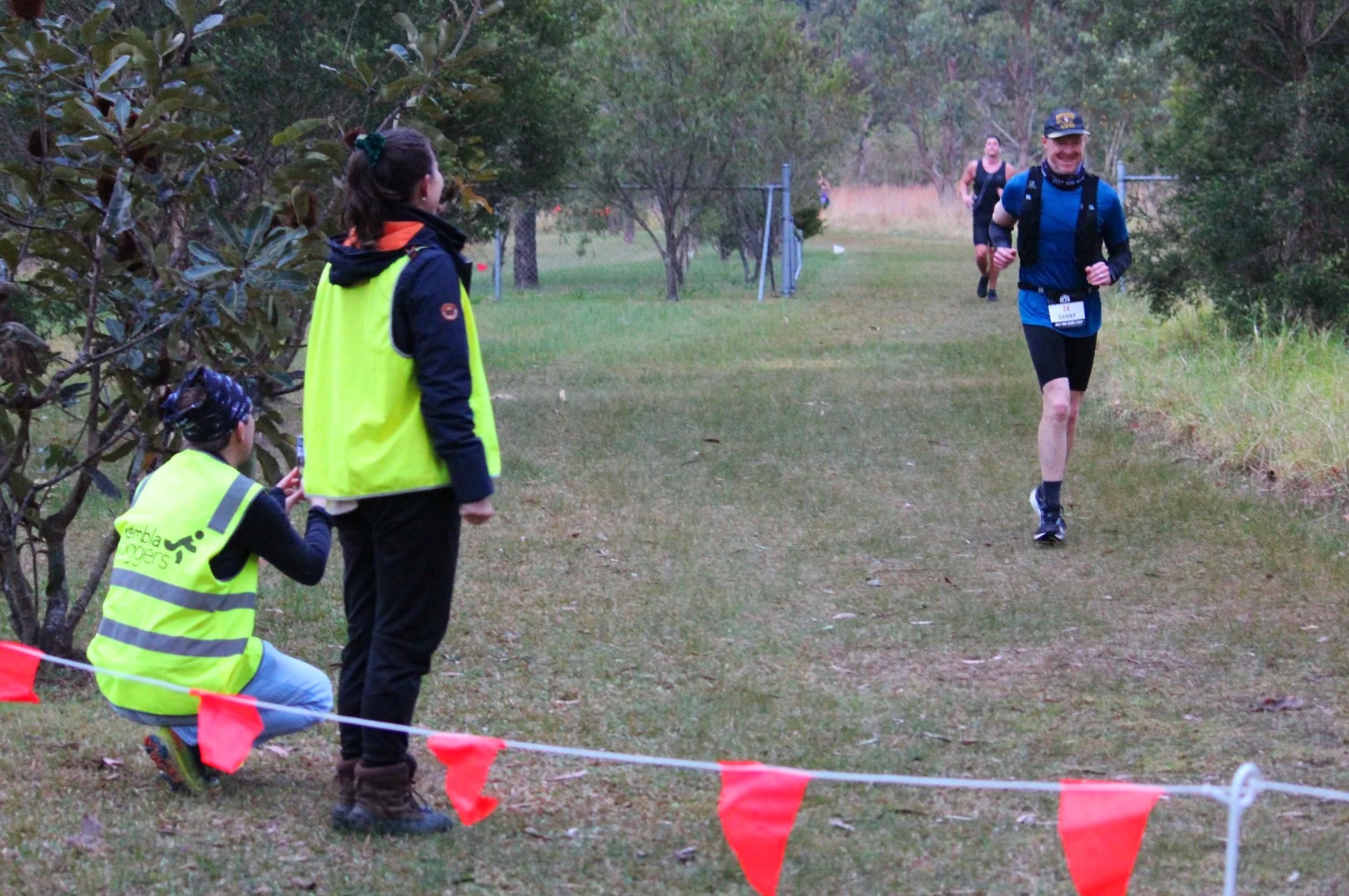 A man running on a grassy trail, wearing a blue shirt, black shorts, and a cap, with a bib number, in a wooded outdoor setting. Spectators in yellow vests are watching from the side.