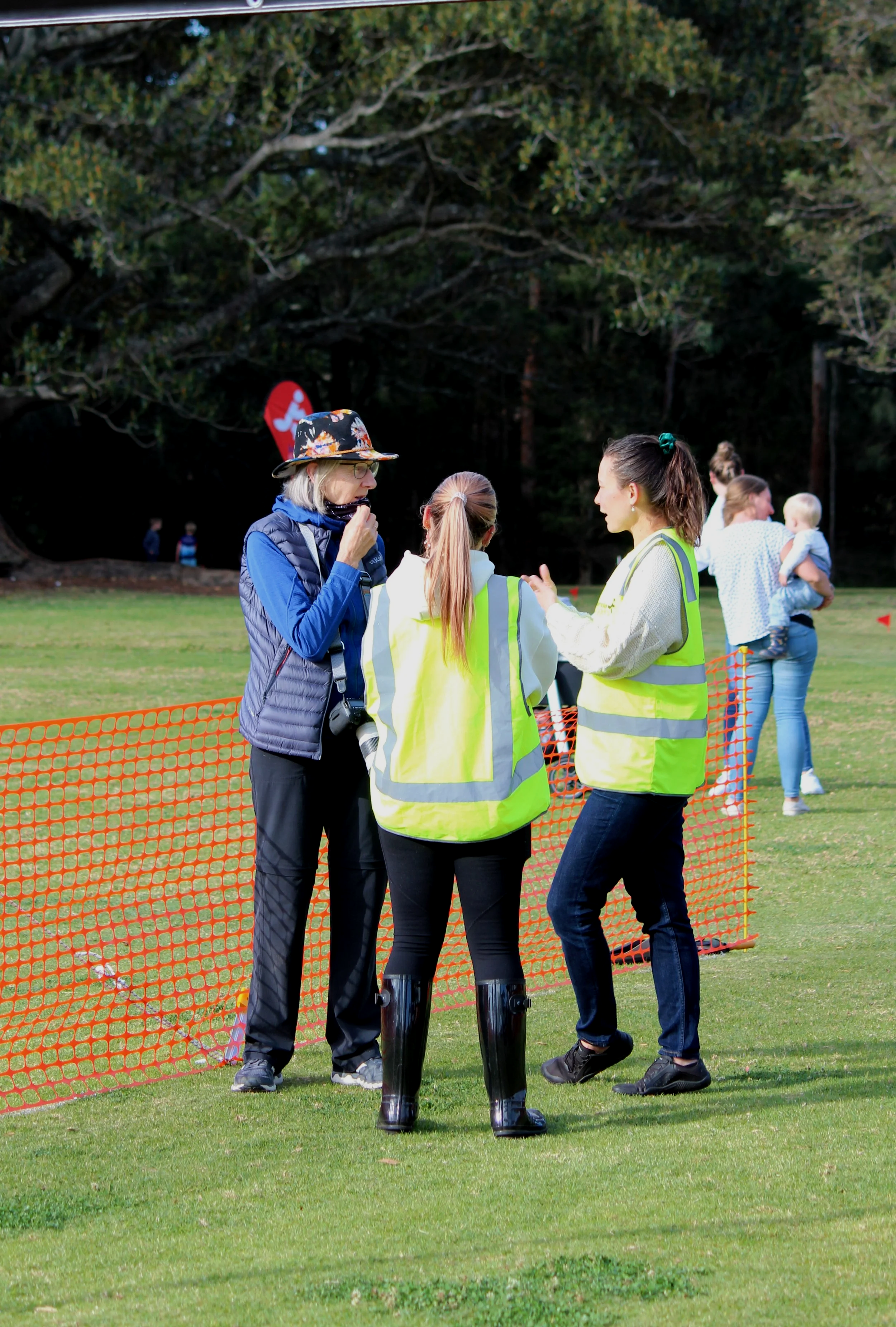 Three women standing and talking on a grassy field near an orange safety barrier, with a forest in the background. One woman wears a blue jacket and a floral hat, another wears a neon yellow vest, and the third woman, also wearing a neon yellow vest,